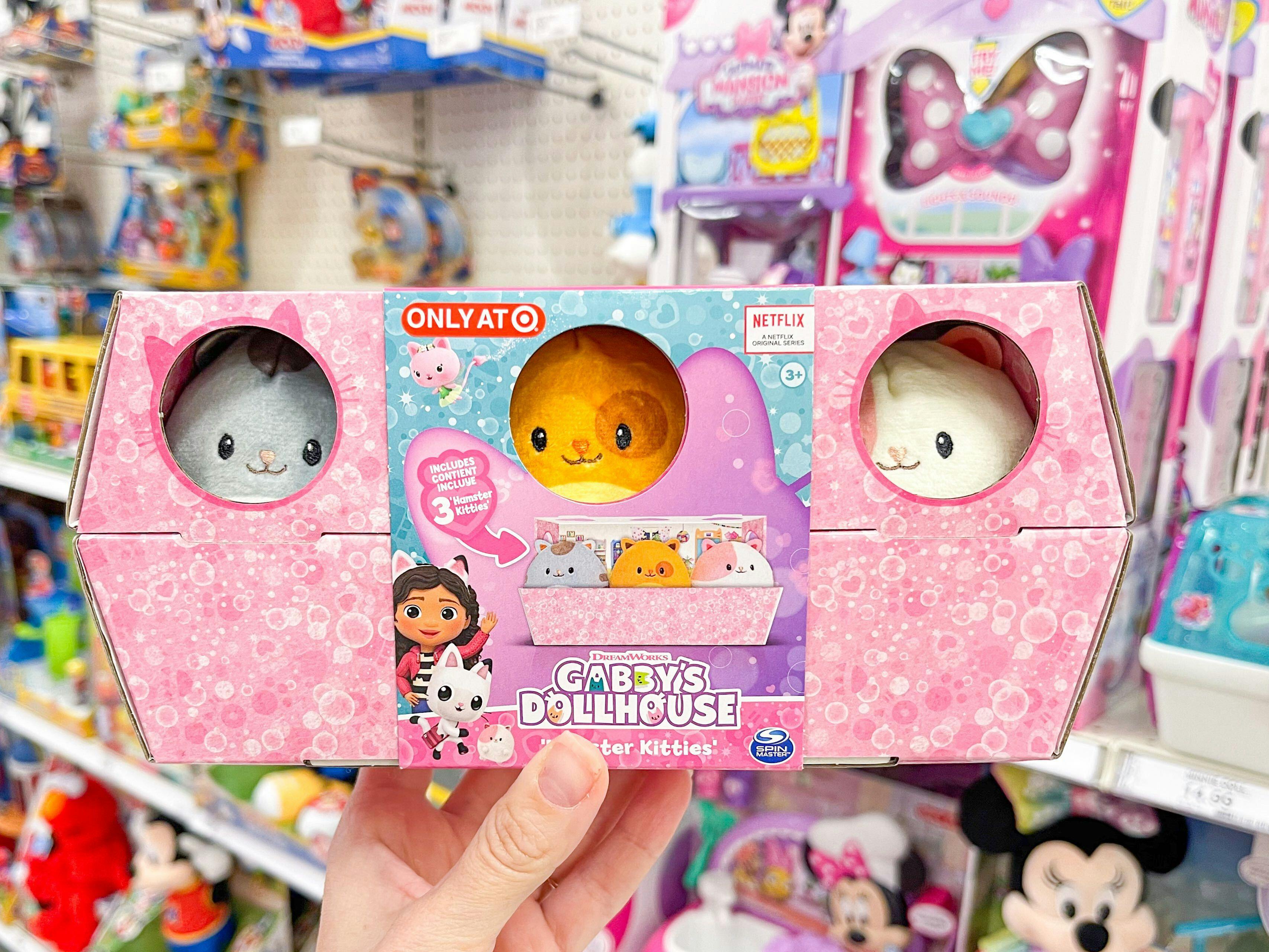 A Gabby's Dollhouse toy held out by hand in front of other toys hanging from store racks.
