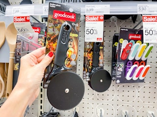 A Goodcook jumbo pizza cutter held out in front of a sale sticker hanging from a store shelf with other kitchen products hanging from a store rack.