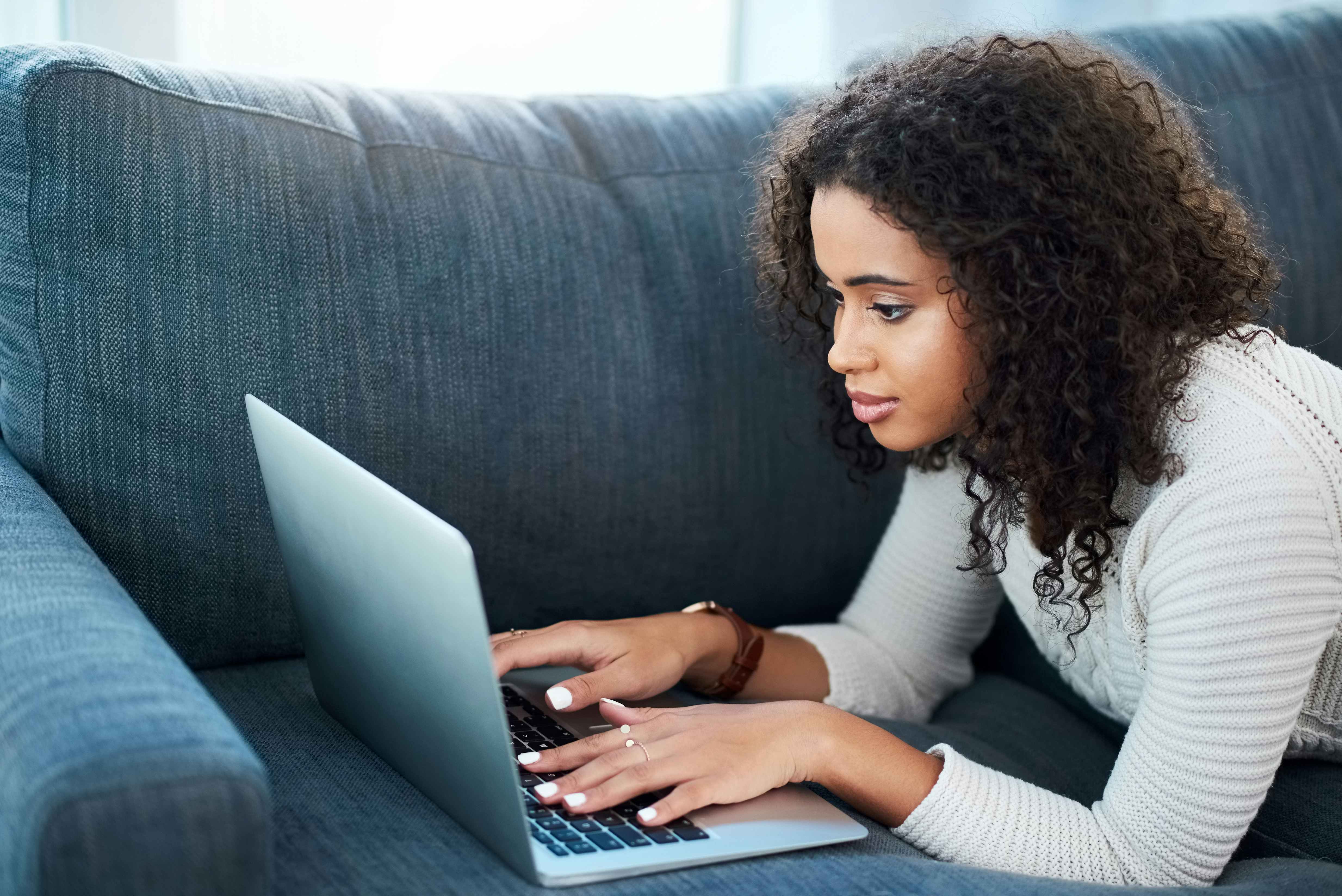 Young woman at her laptop typing while laying on the couch
