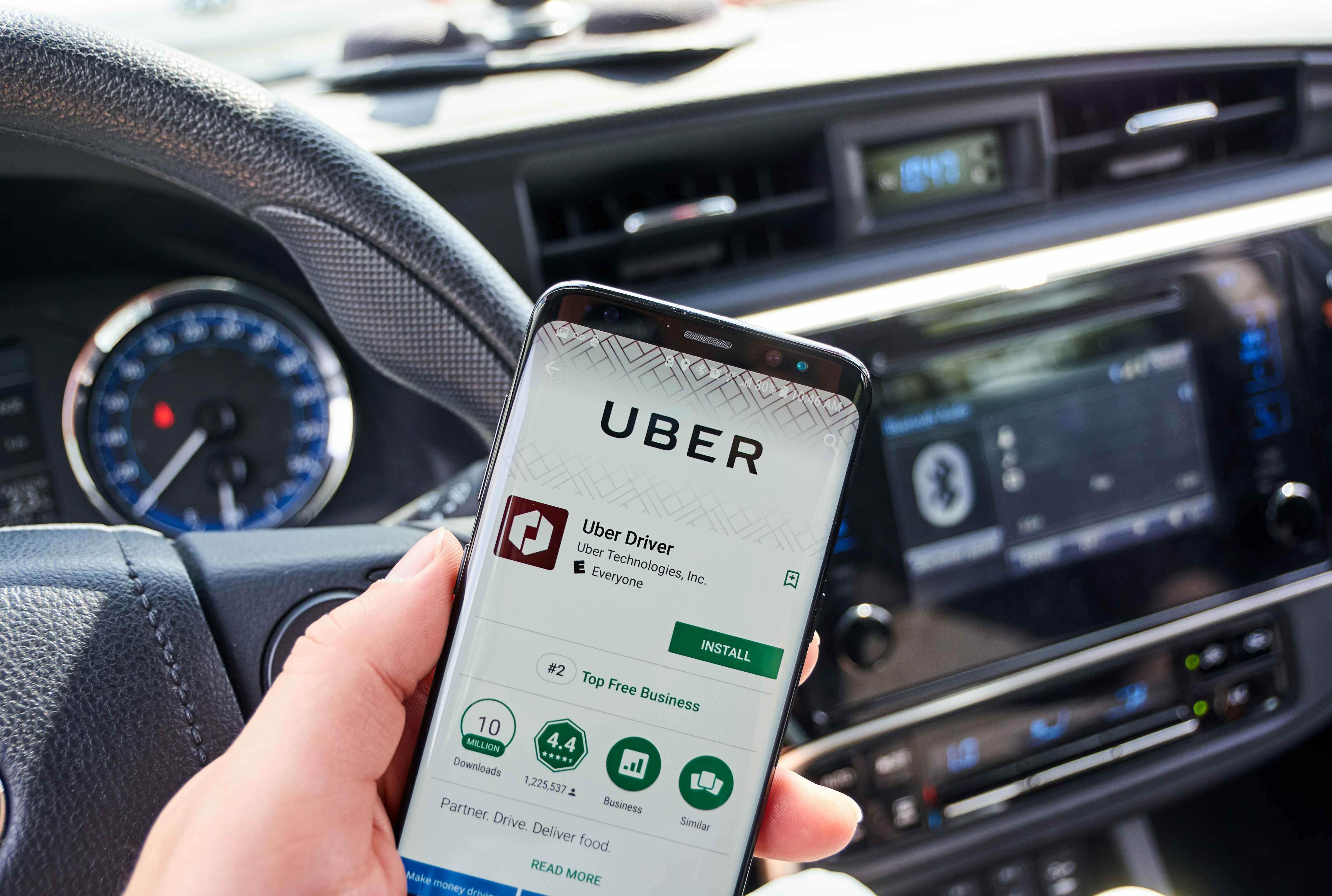 A person in the drivers seat of a car holding a phone with the uber driver app download page on the screen.