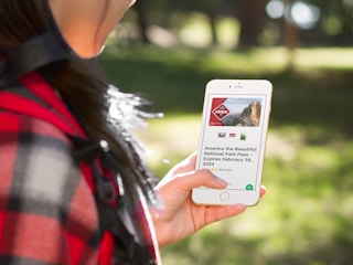 A woman holding up her smartphone and looking at an american the beautiful pass for $80 on usparkpasses.com