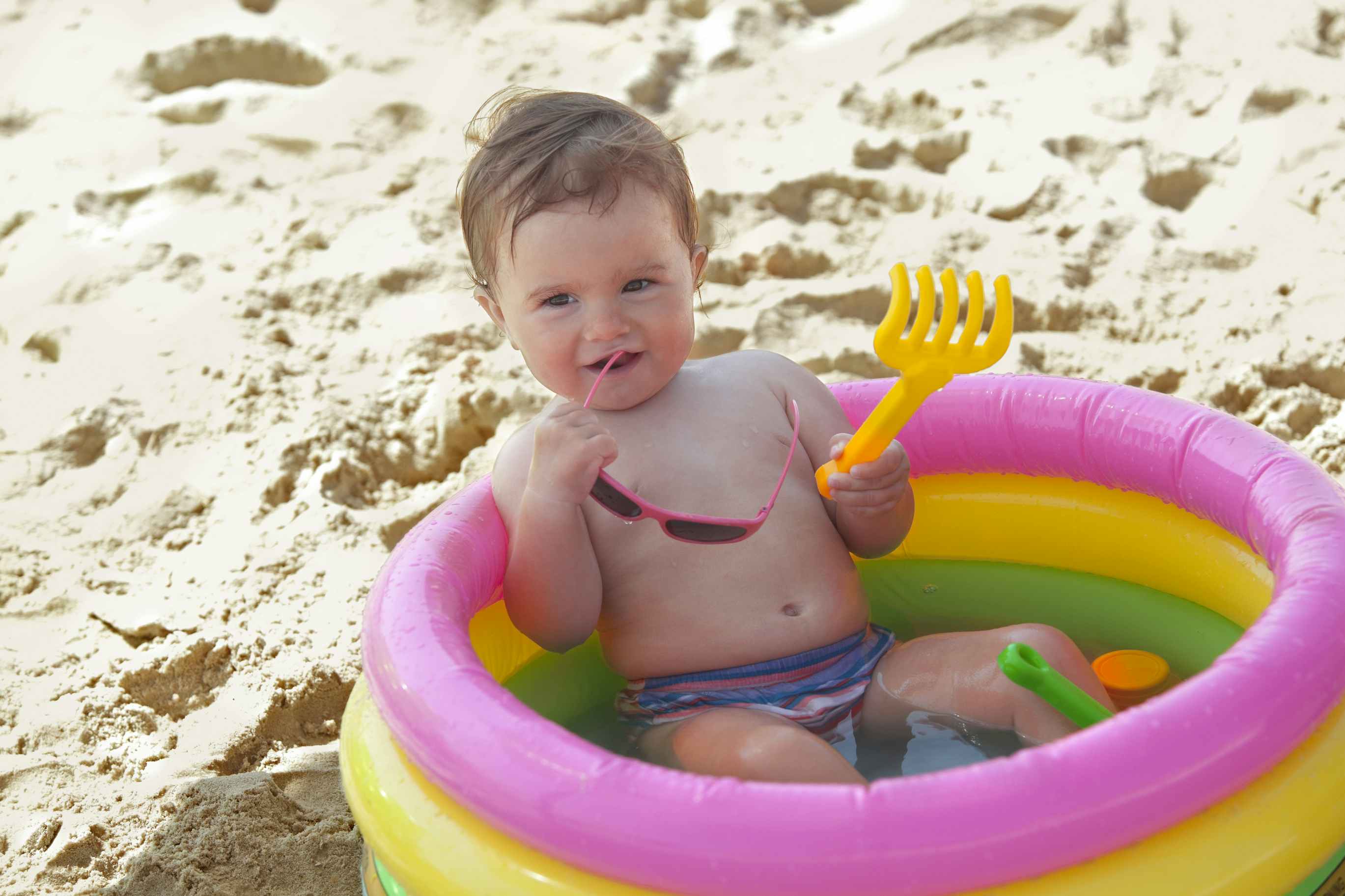 Baby sits inside a kiddie pool at the beach. The baby is holding a plastic toy and a pair of sunglasses