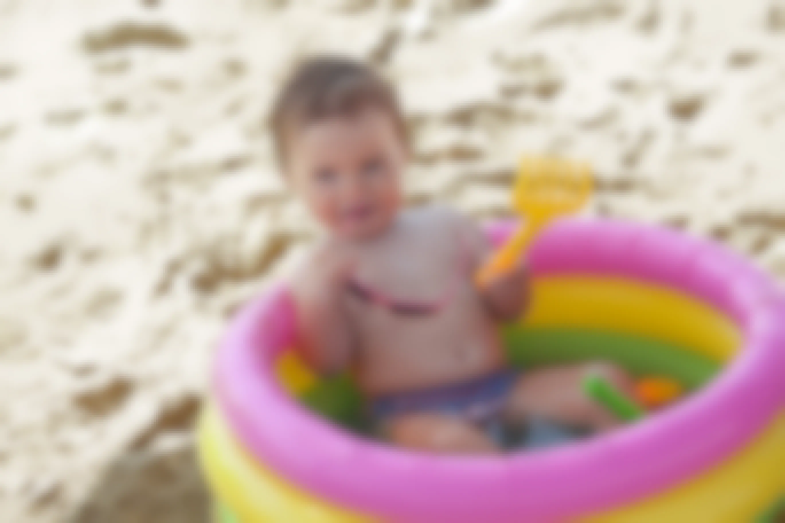 Baby sits inside a kiddie pool at the beach. The baby is holding a plastic toy and a pair of sunglasses