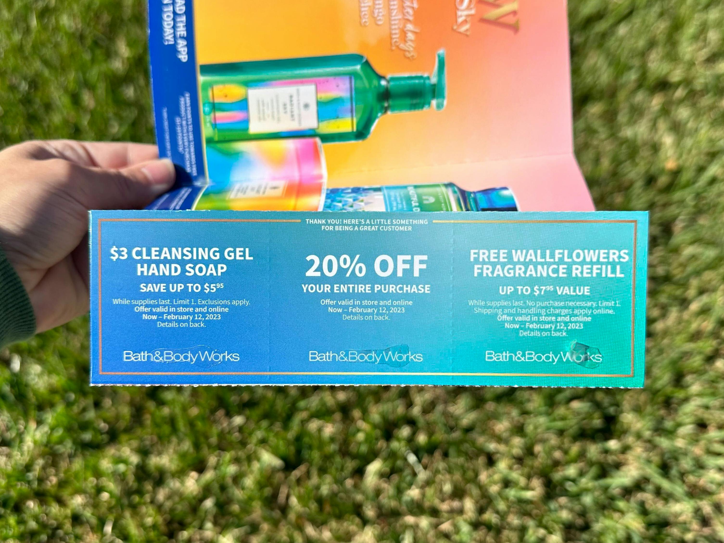 Someone holding a Bath & Body Works mailer coupon book in front of their lawn