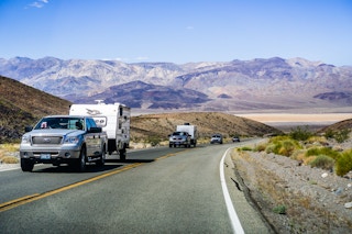 Two trucks towing RV campers driving up a hill with scenic mountains in the background