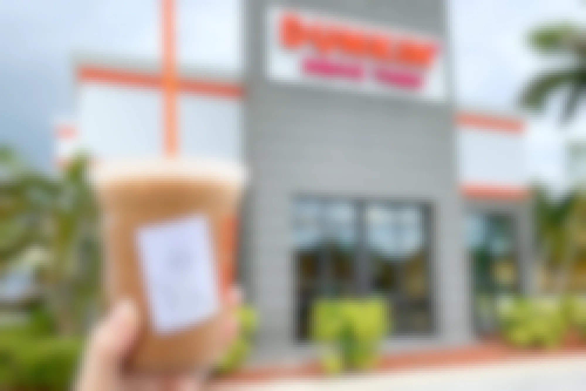 A person's hand holding up a specialty iced coffee outside of a Dunkin restaurant.