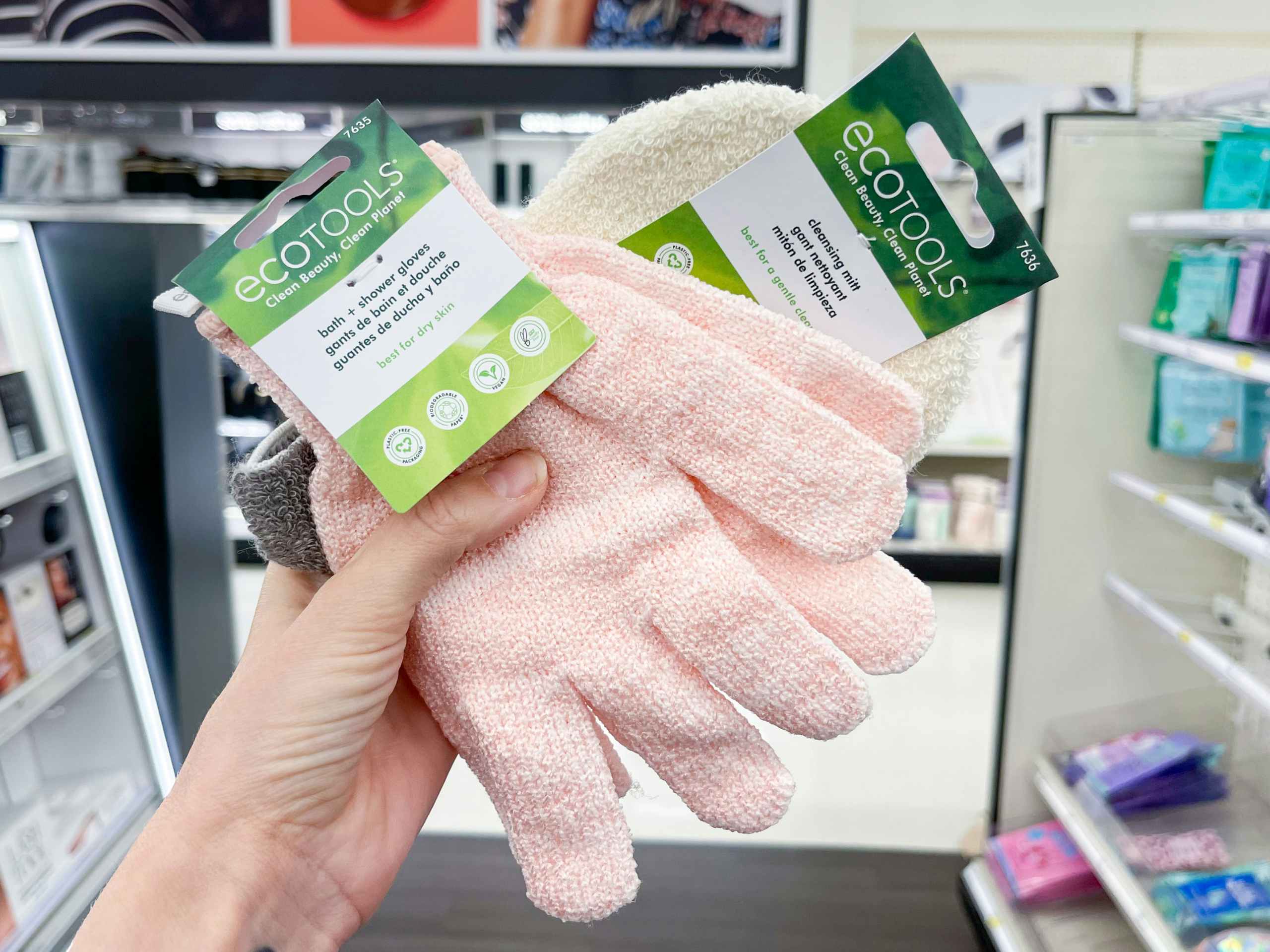 A couple of ecoTools bath gloves held out by hand in front of a store aisle.