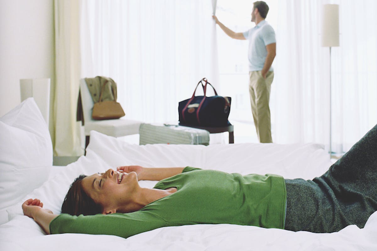 Woman lays on a hotel bed while another person is looking outside from the hotel window