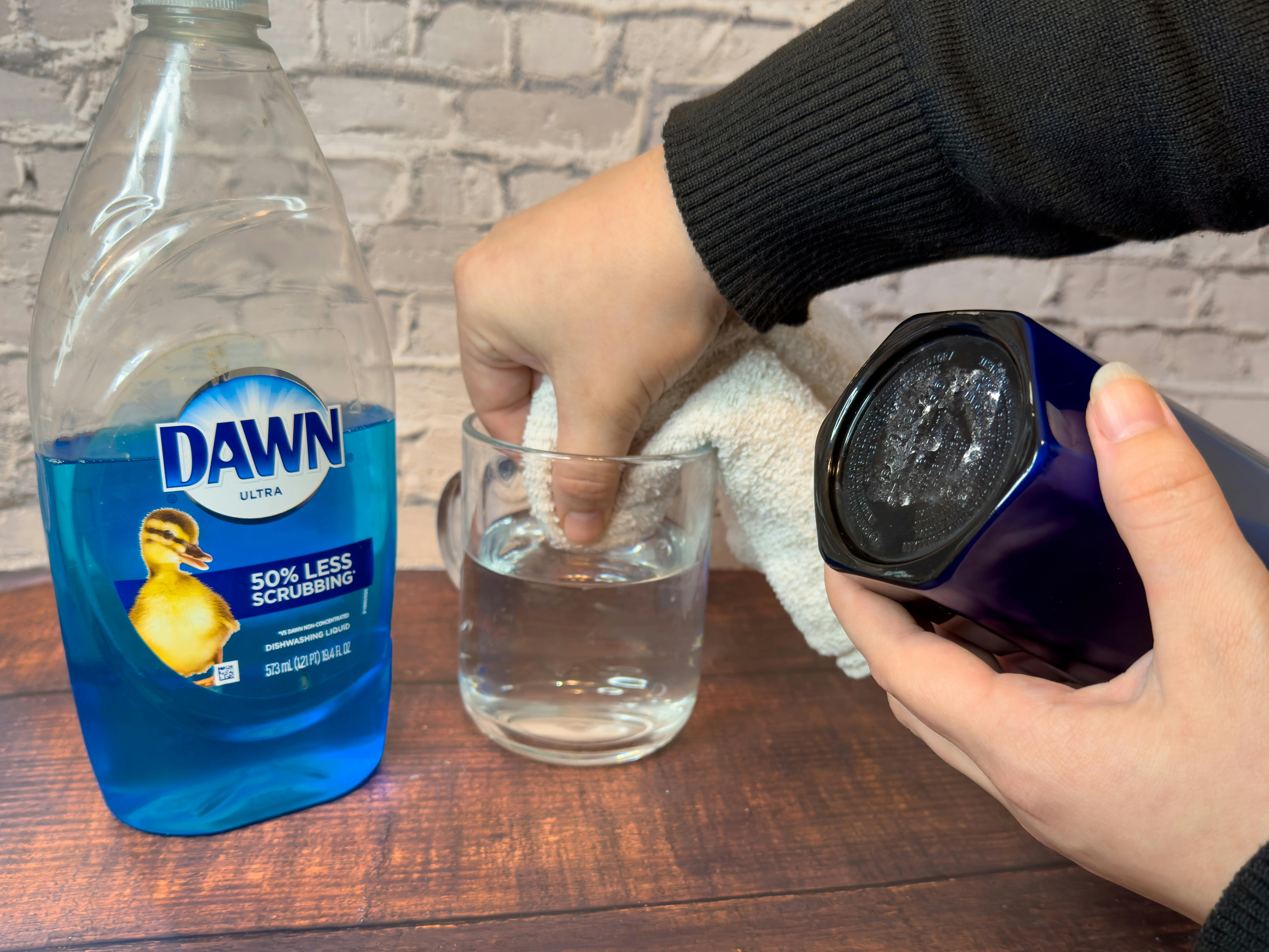 Someone dipping a rag into a glass of warm water next to a bottle of Dawn dish soap and holding a Starbucks cup showing the sticker residue left on the bottom