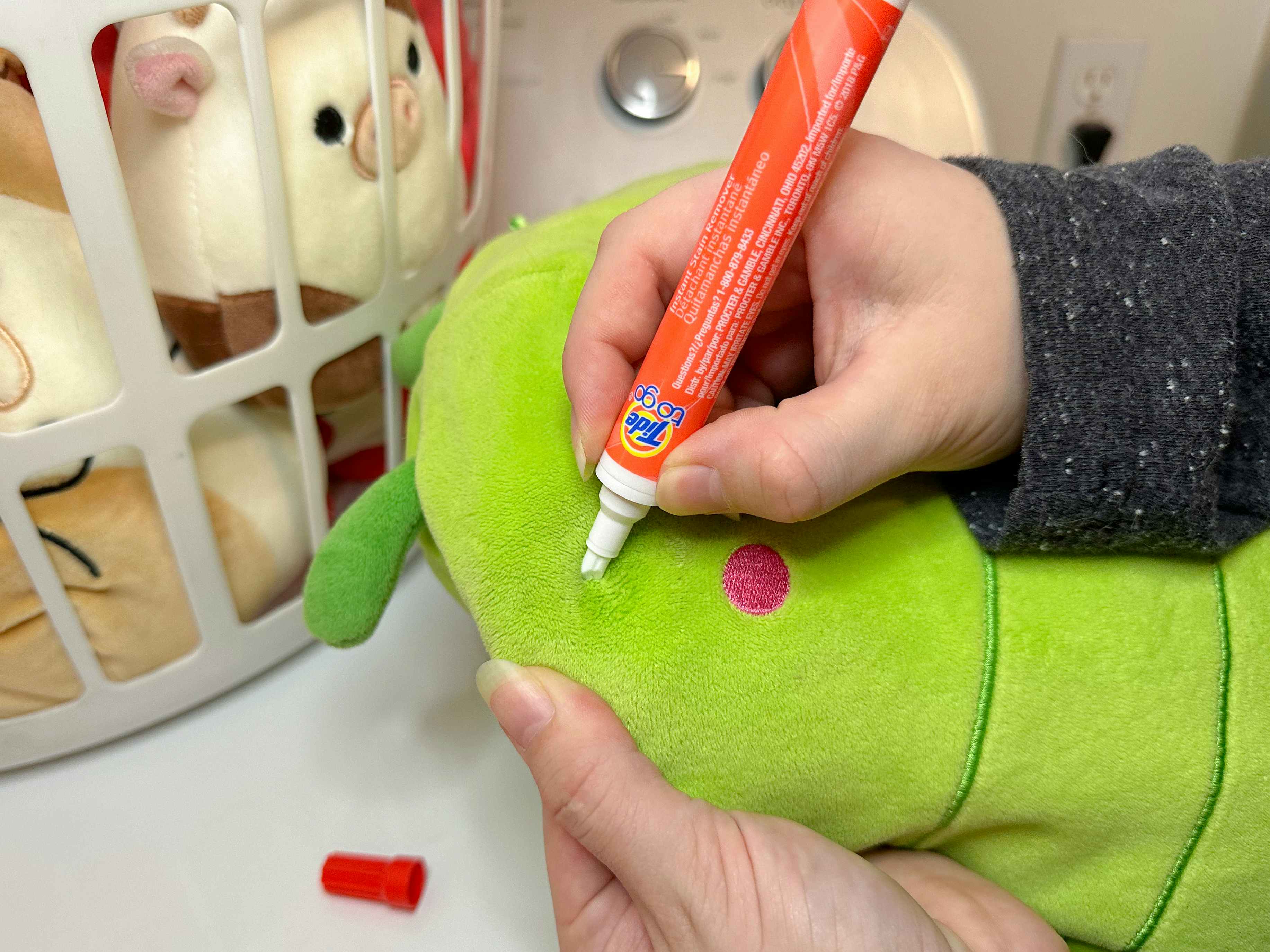 Someone using a Tide pen on a Squishmallow next to a laundry basket full of Squishmallows