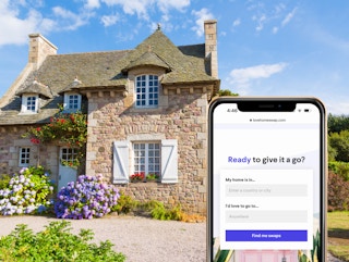 A close up shot of a smartphone showing the loveswap.com house swapping site in front of a charming country cottage