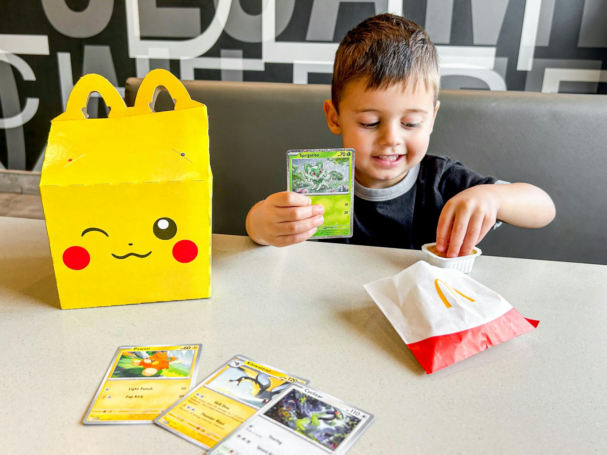 a child eating a chicken nugget while sitting with the pokemon happy meal at mcdonalds