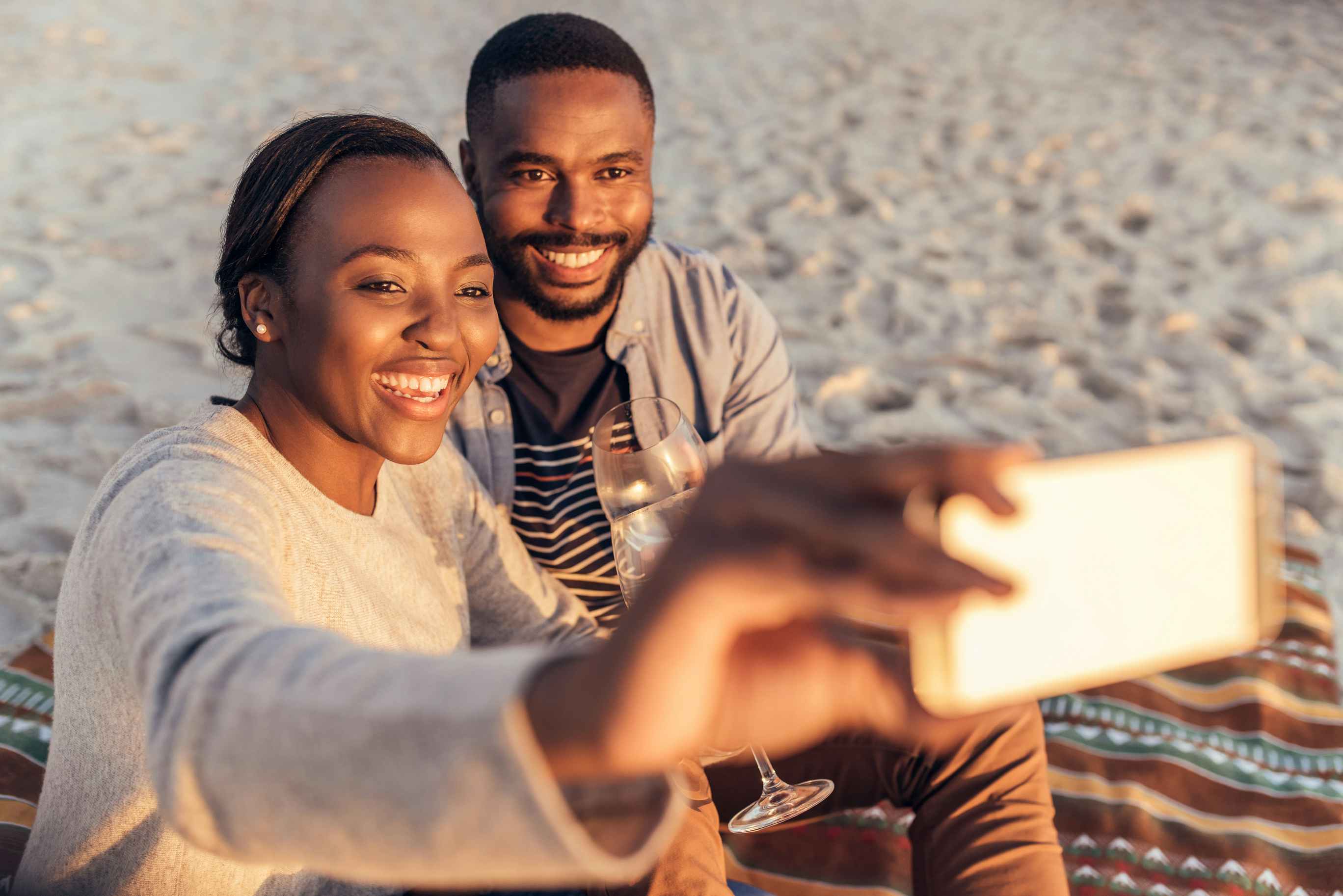 Two people sitting together on a blanket at the beach and taking a selfie