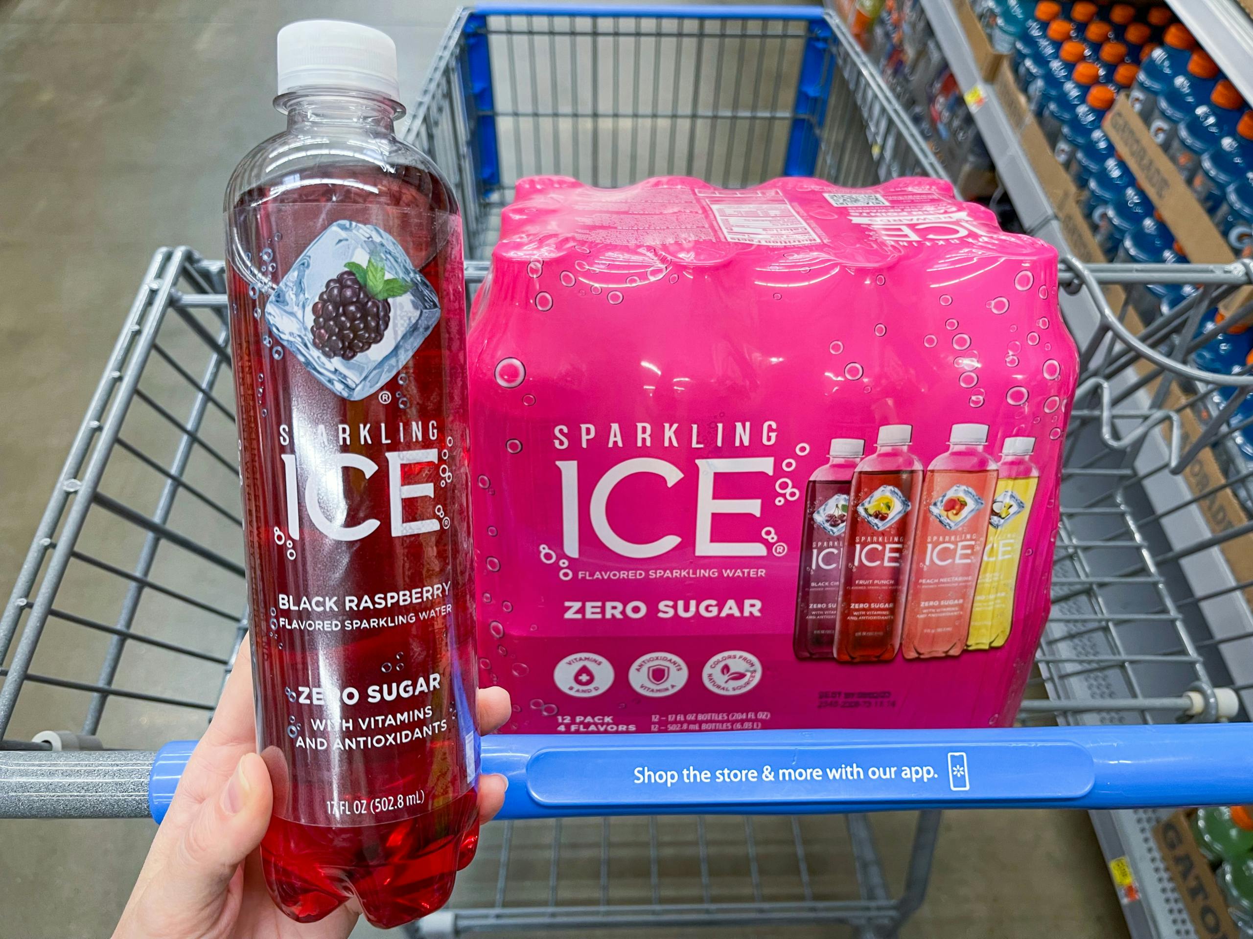 A Sparkling Ice Black Raspberry held out by hand in front of a Sparkling Ice variety package sitting in a store cart.