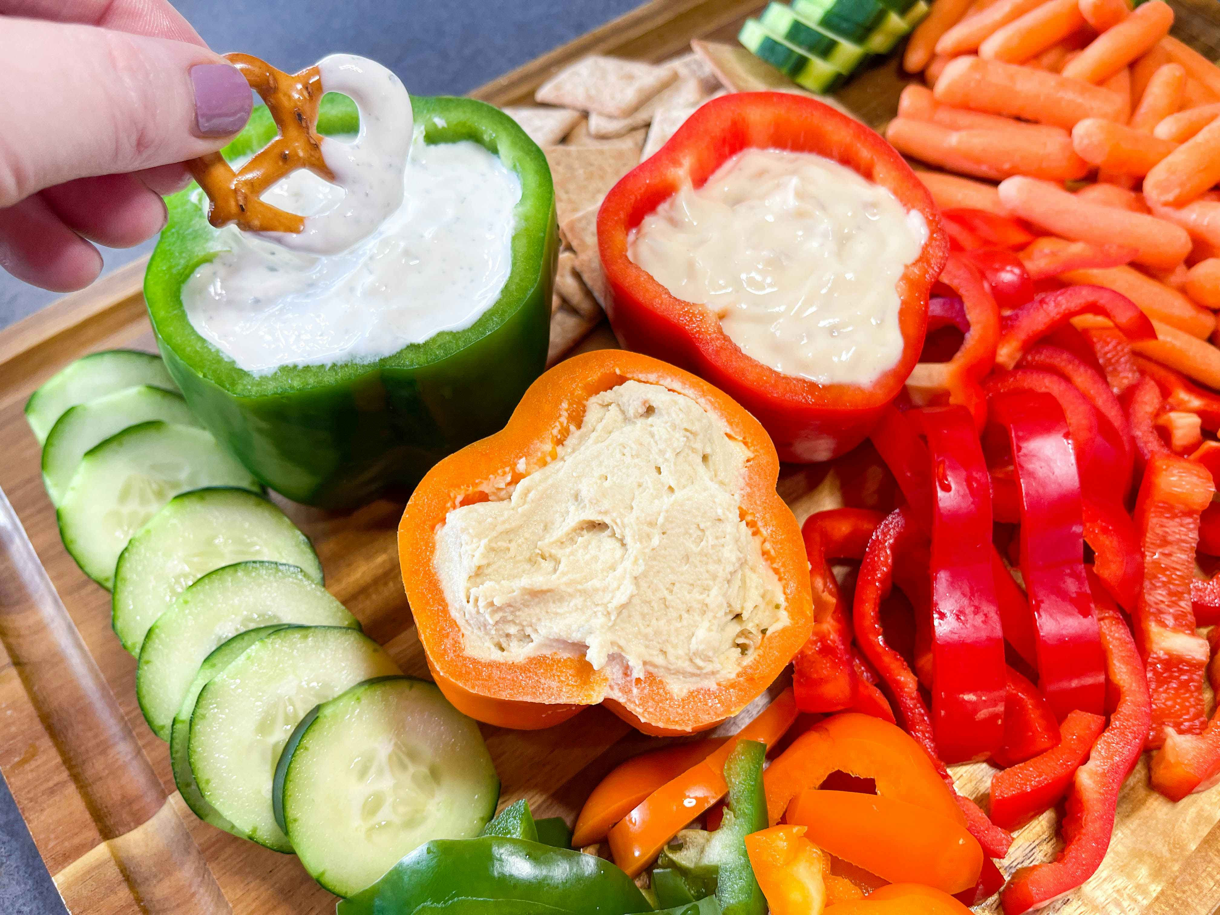 a hand dipping a pretzel into dip from a bell pepper filled with dips on a veggie tray 