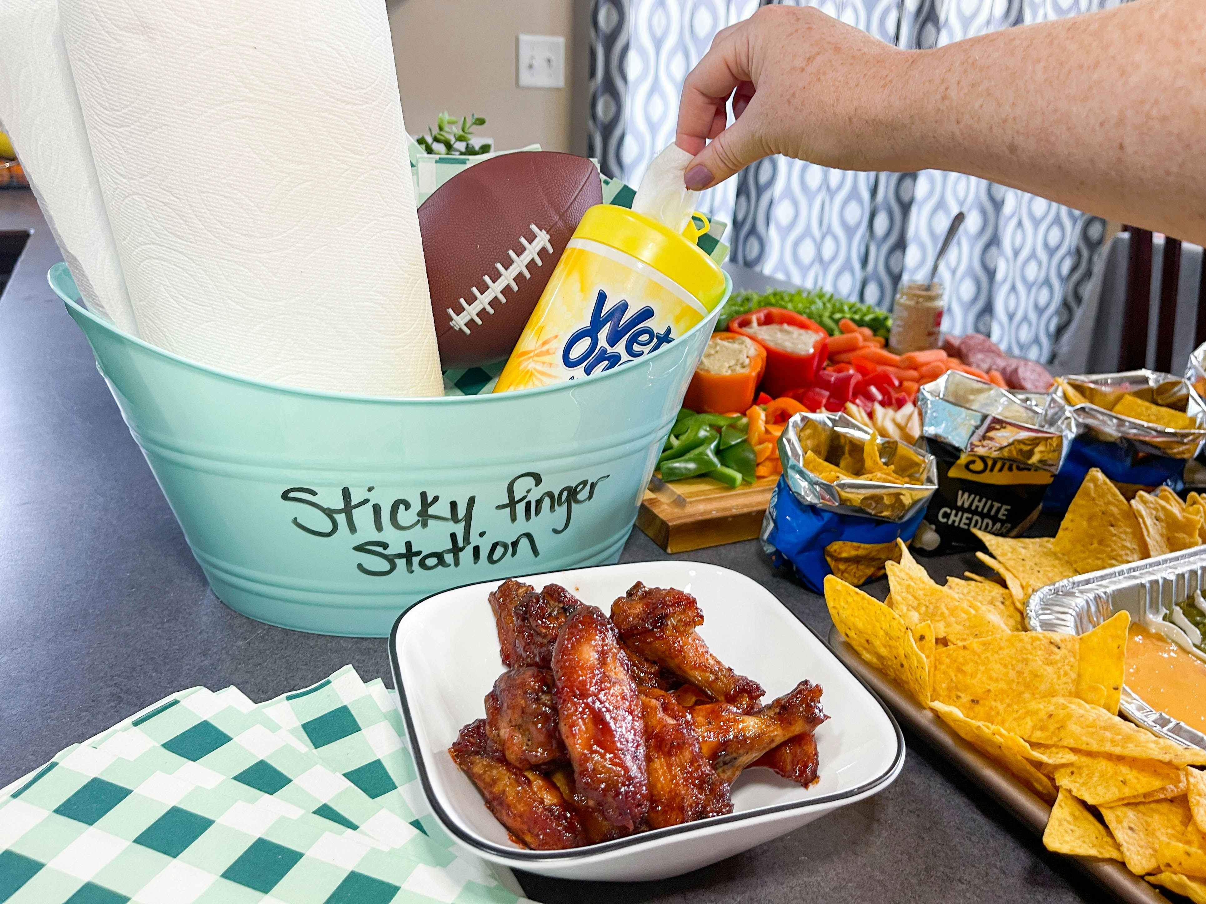 a diy sticky finger station bucket with wet wipes, football, and paper towels, next to wings on a counter 