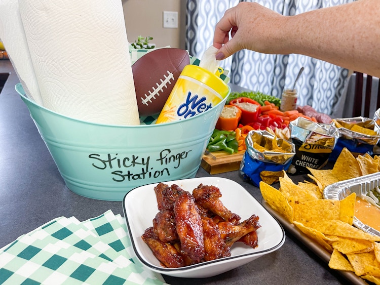 a diy sticky finger station bucket with wet wipes, football, and paper towels, next to wings on a counter