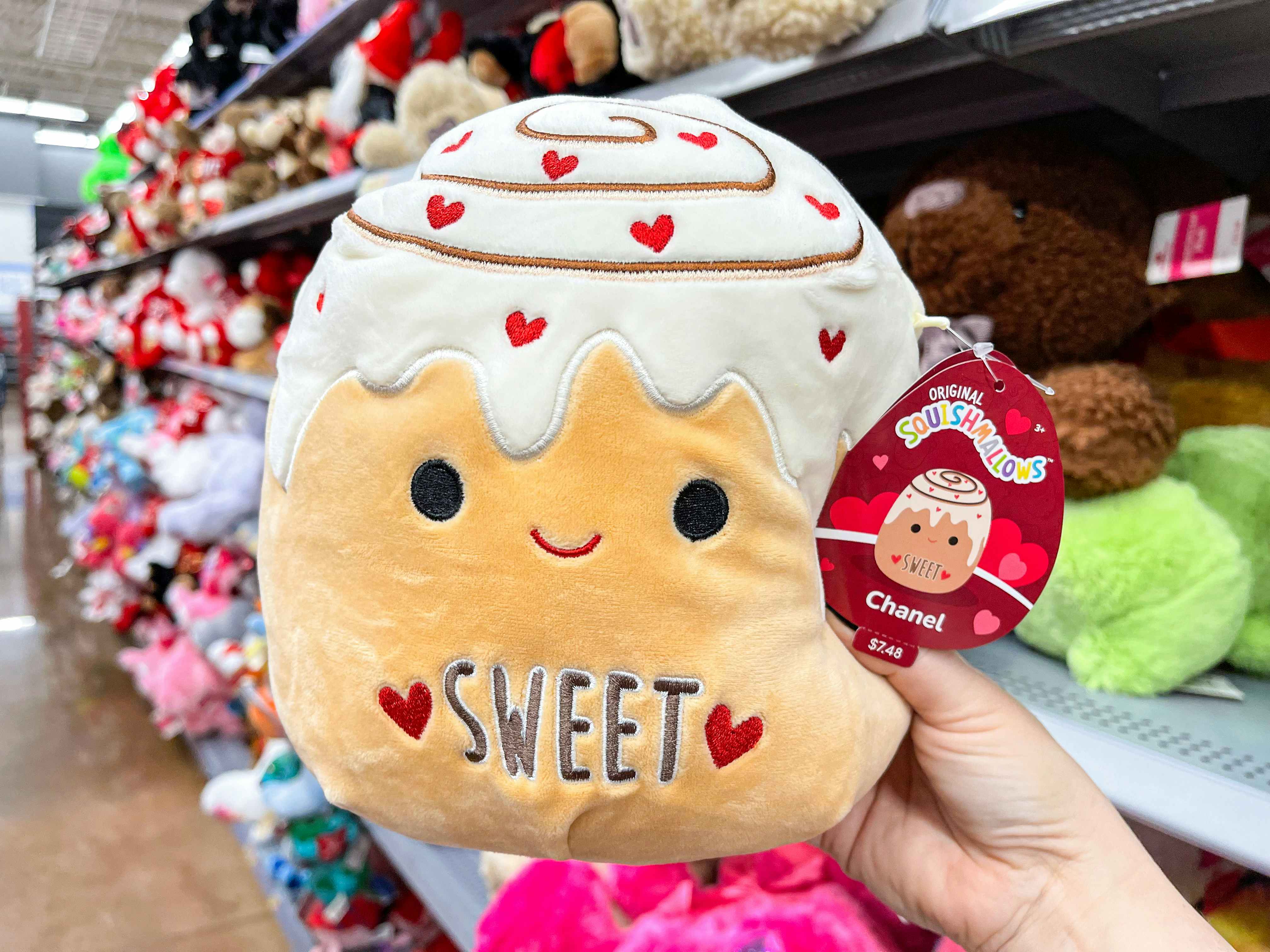 Someone holding up a Cinnamon Roll Squishmallow that says "Sweet" on the tummy.