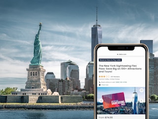 A view of the statue of liberty and downtown manhattan NYC behind a smartphone showing an NYC sightseeing pass on Viator.com