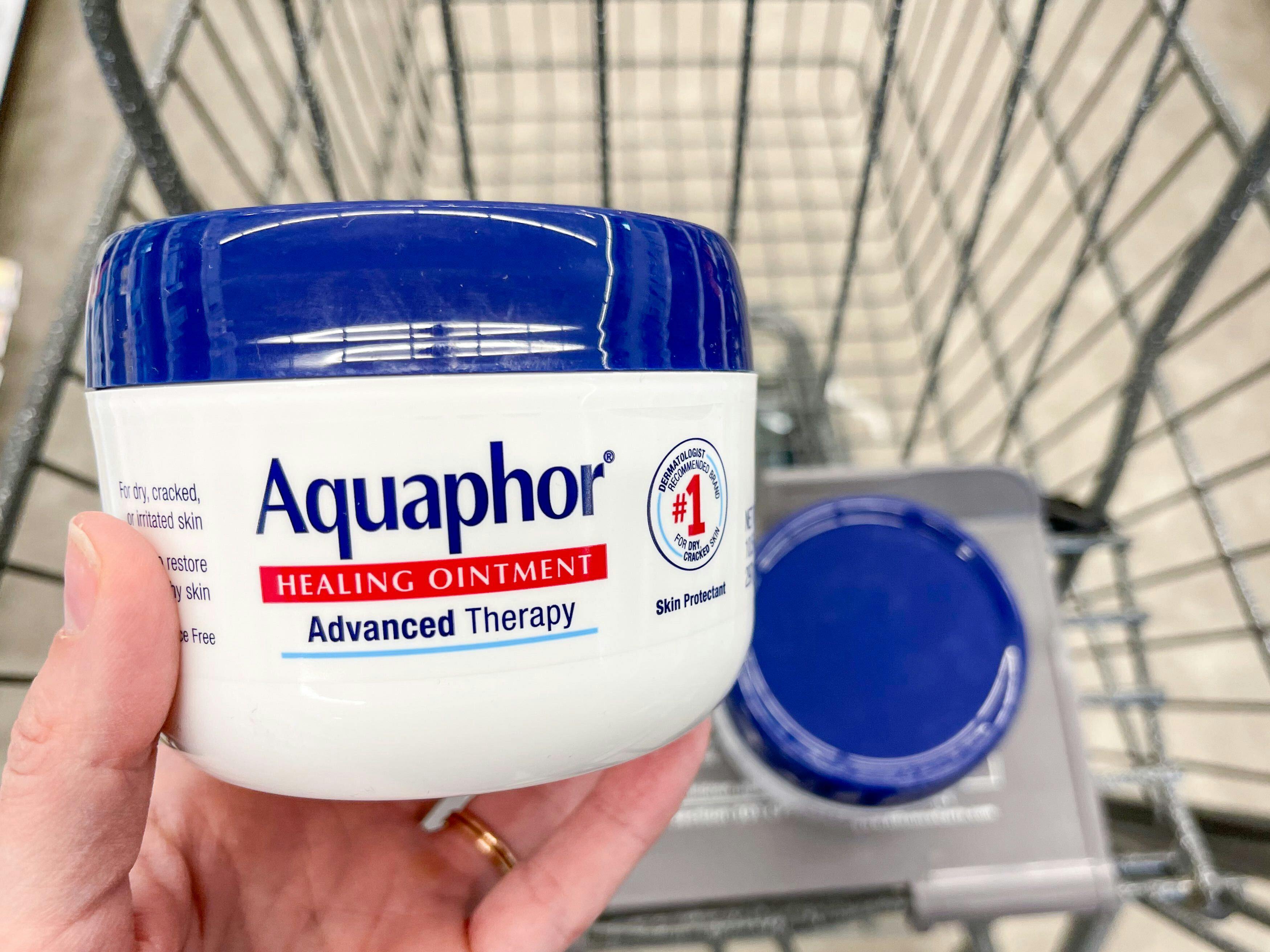 An Aquaphor jar held out by hand in front of another Aquaphor jar sitting in a store cart