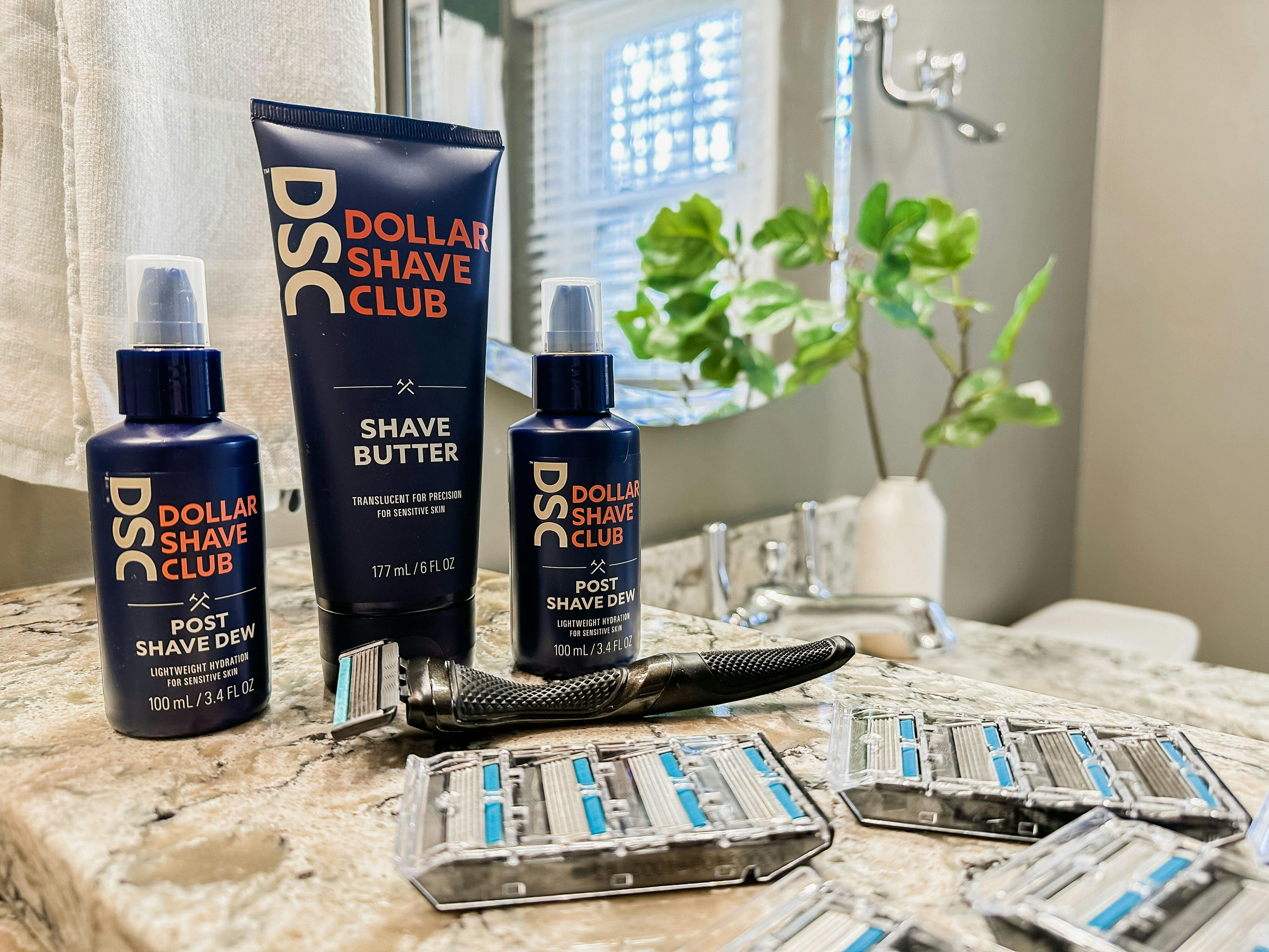 The Dollar Shave Club Shave Butter and two bottles of DSC Post Shave Dew lined up on the counter behind a handful of DSC razor cartridges