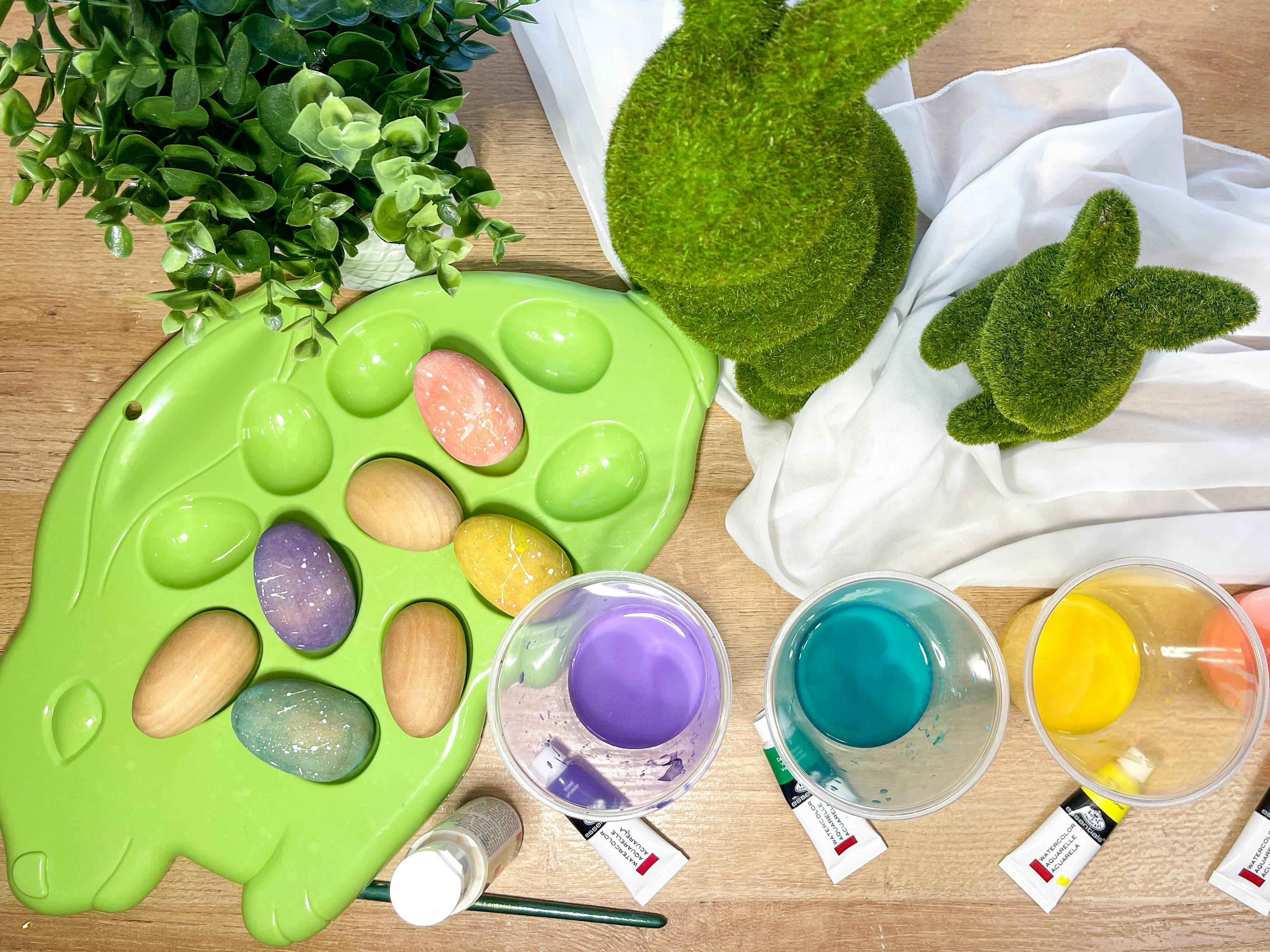 Watercolor painted wood eggs on a table with easter decor and cups of watercolor paint