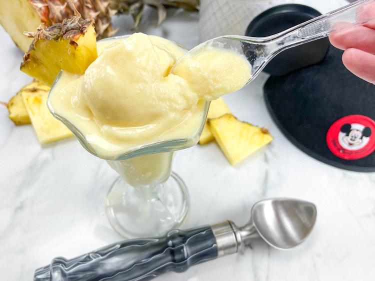 homemade dole whip in an ice cream cup on a counter next to a Mickey ears hat and a pineapple