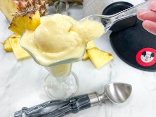 homemade dole whip in an ice cream cup on a counter next to a Mickey ears hat and a pineapple