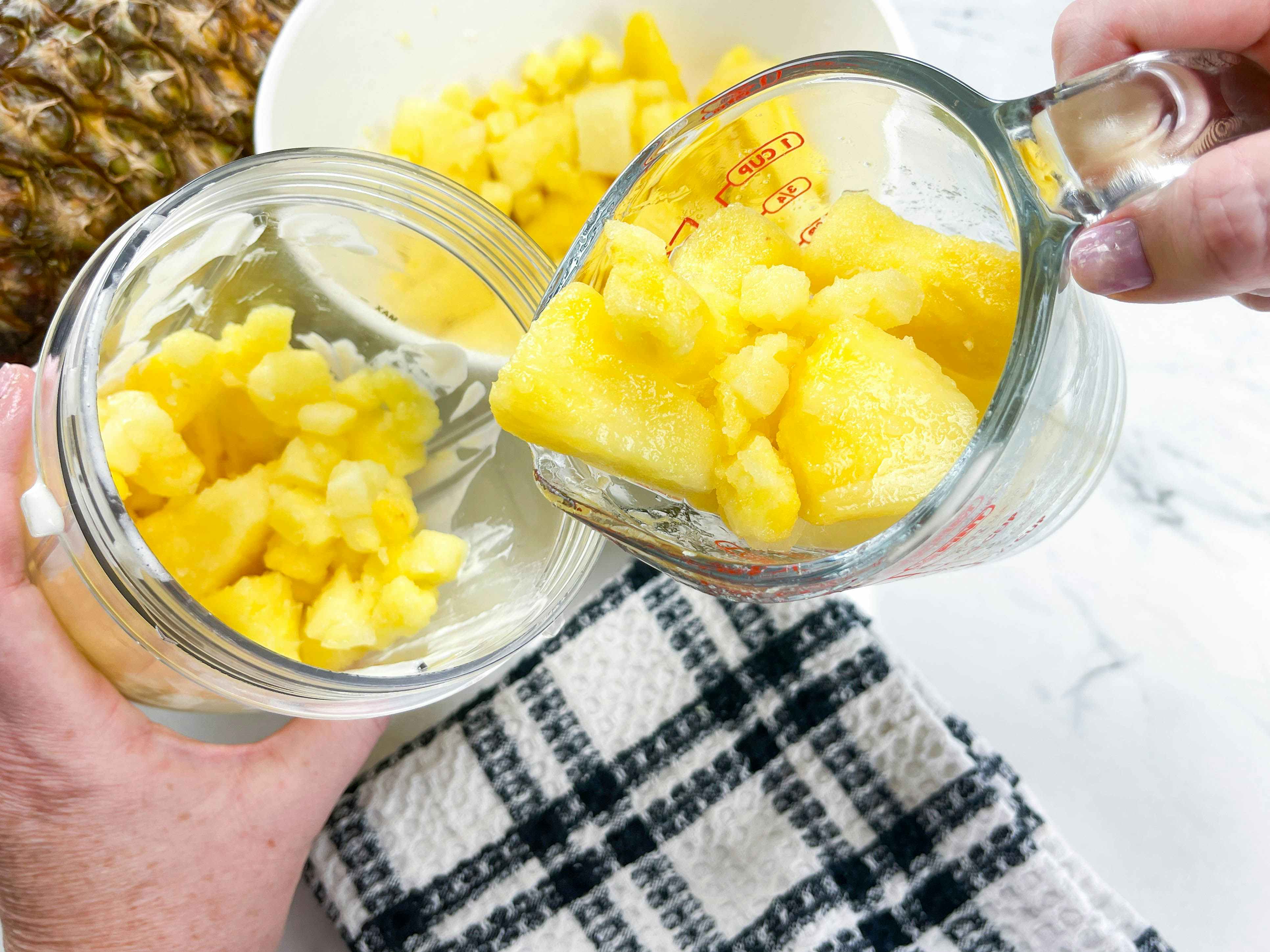 a person pouring a measuring cup full of frozen pineapple into a blender cup 