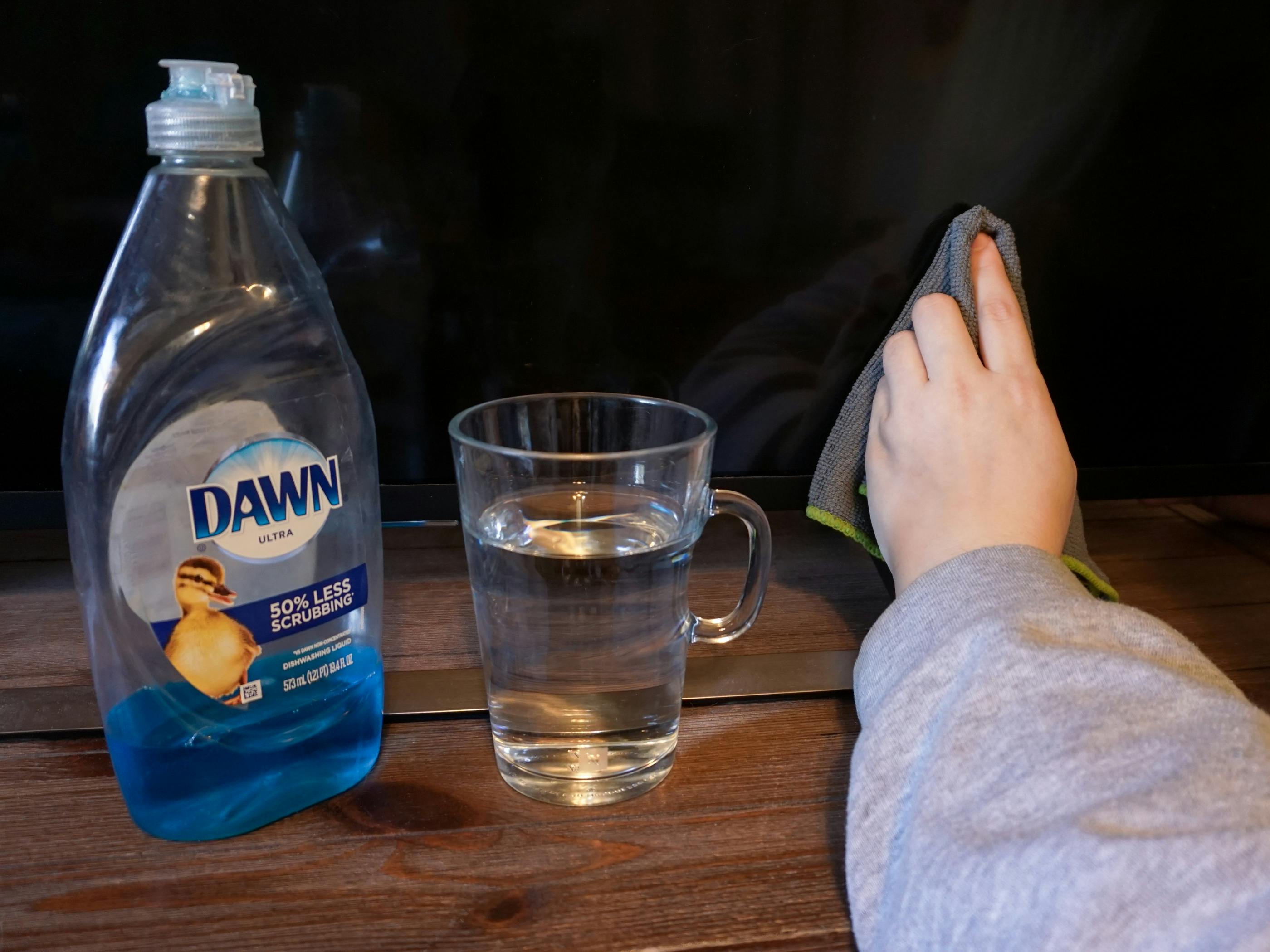 Someone using Dawn dish soap diluted in water to get a smear off of their TV with the bottle of dawn and a glass of water on the TV stand 
