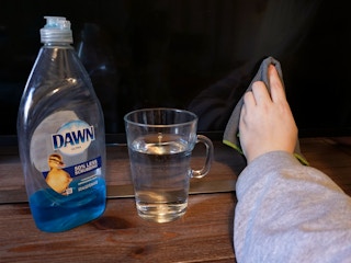 Someone using Dawn dish soap diluted in water to get a smear off of their TV with the bottle of dawn and a glass of water on the TV stand