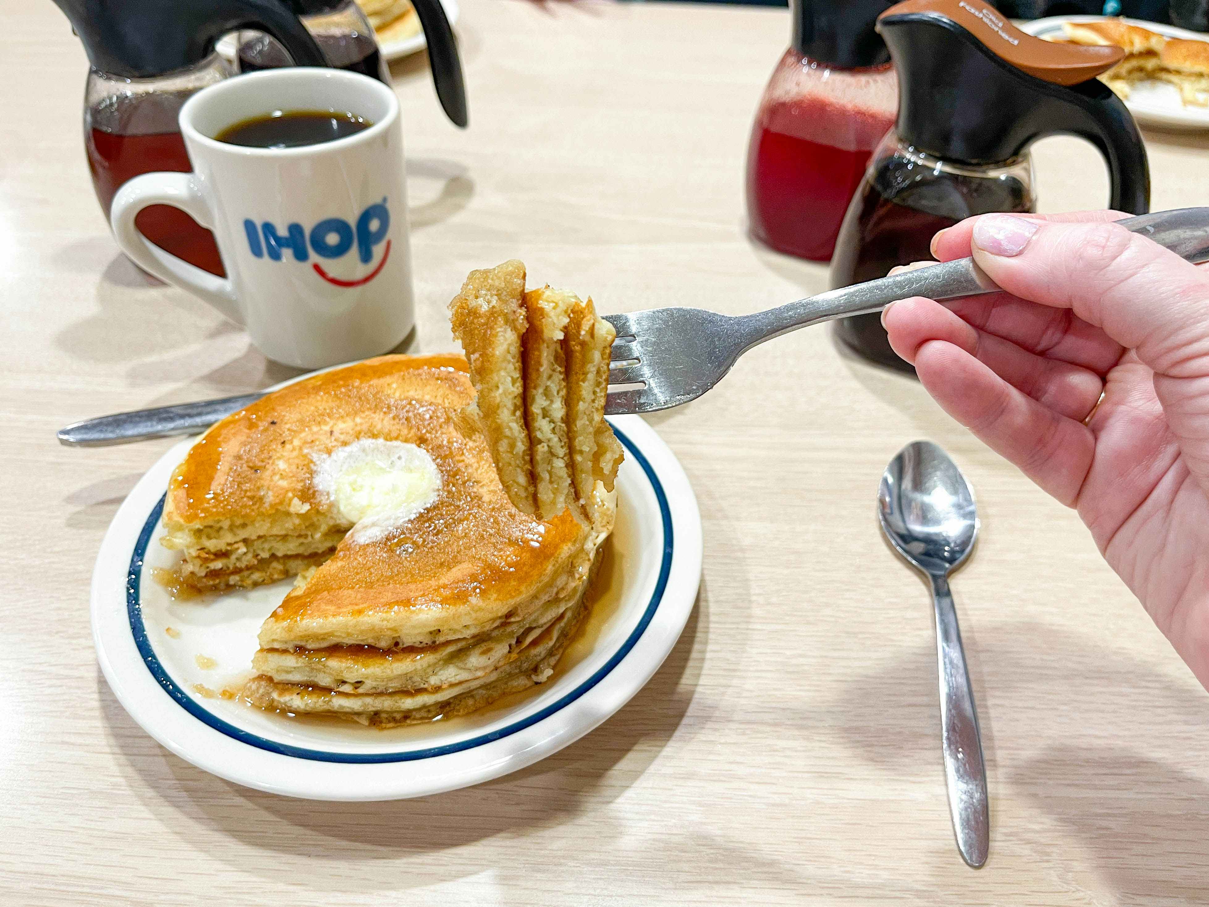a plate with pancakes on a table with syrups and a cup of coffee at ihop with a fork full of pancakes