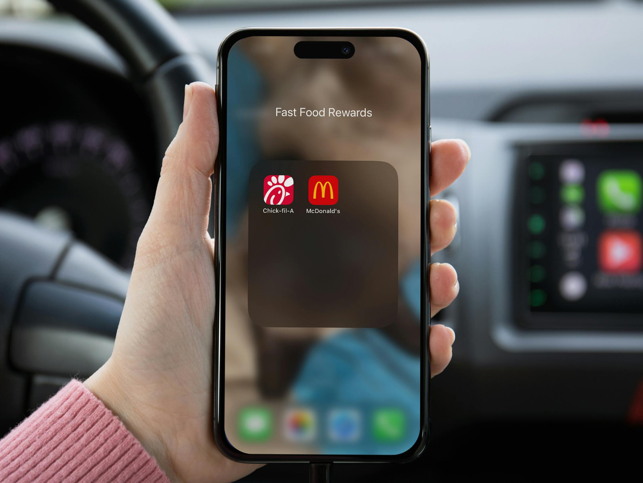 chick-fil-a vs. mcdonalds - Someone sitting in a car, holding a phone displaying an app folder labeled "Fast Food Rewards" with the McDonalds and Chick-fil-A apps organized into it