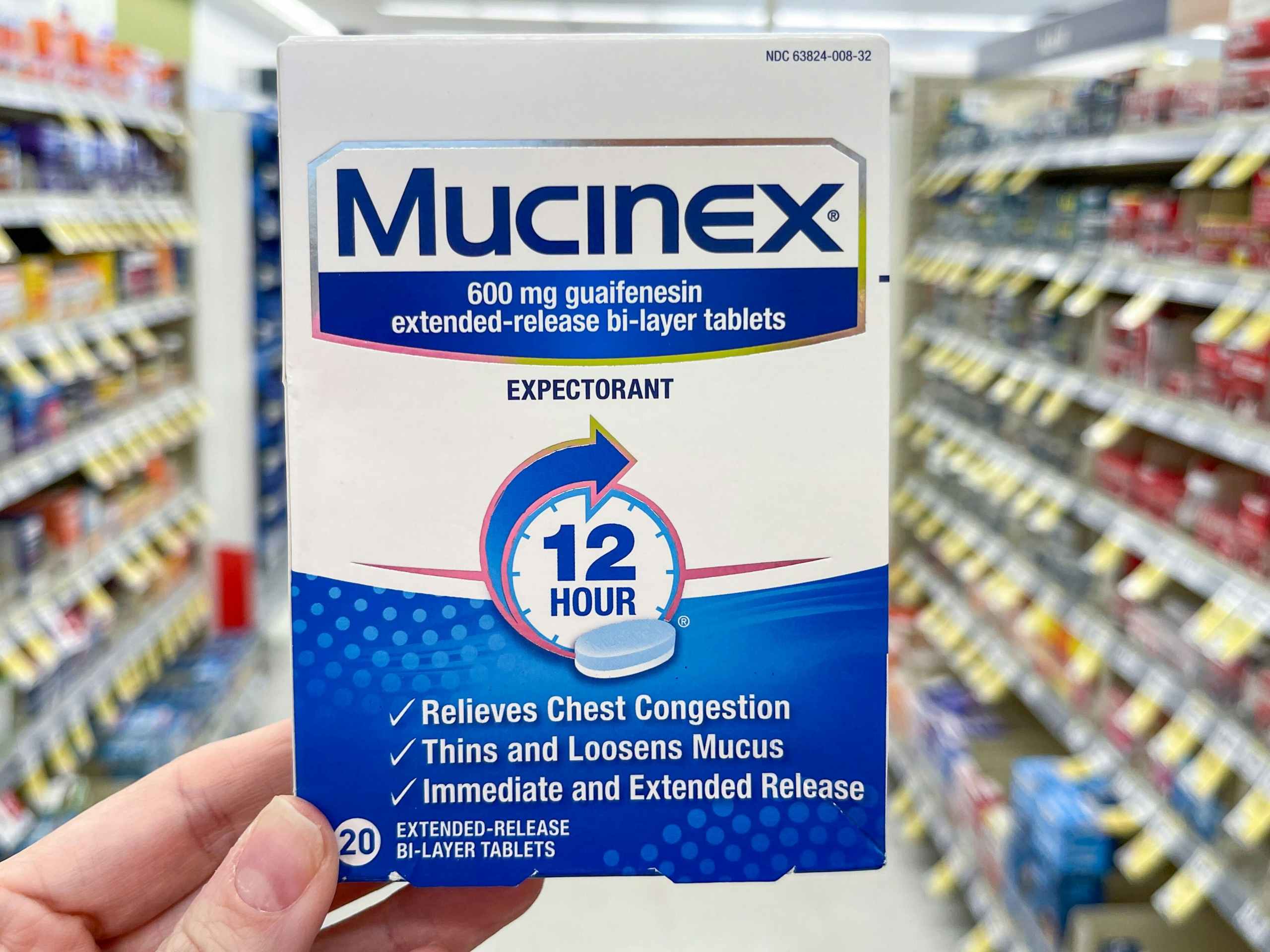 A box of Mucinex Extended Release tablets held out by hand in front of a store aisle.