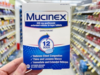 A box of Mucinex Extended Release tablets held out by hand in front of a store aisle.