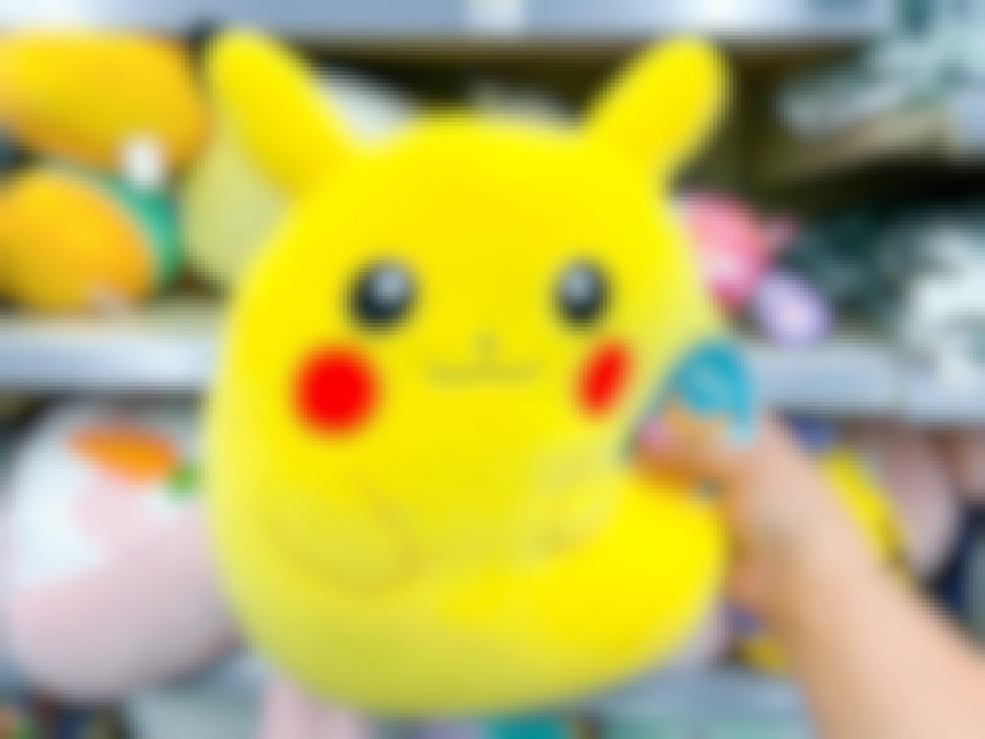 Someone holding a Pikachu Squishmallow in front of some shelves