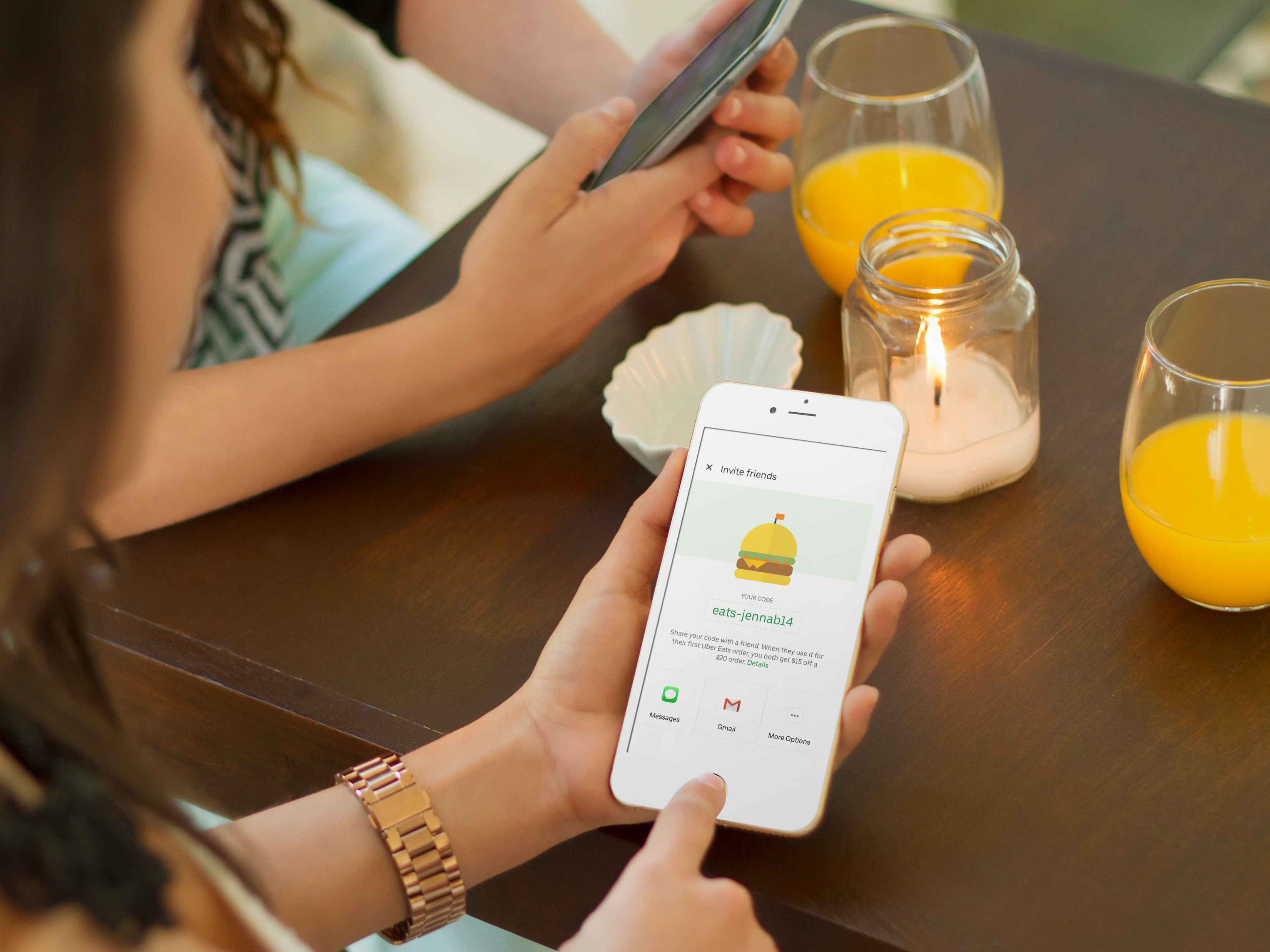 two friends drinking cocktails at a coffee table and one is holding her phone and sending her uber eats referral code to her friend