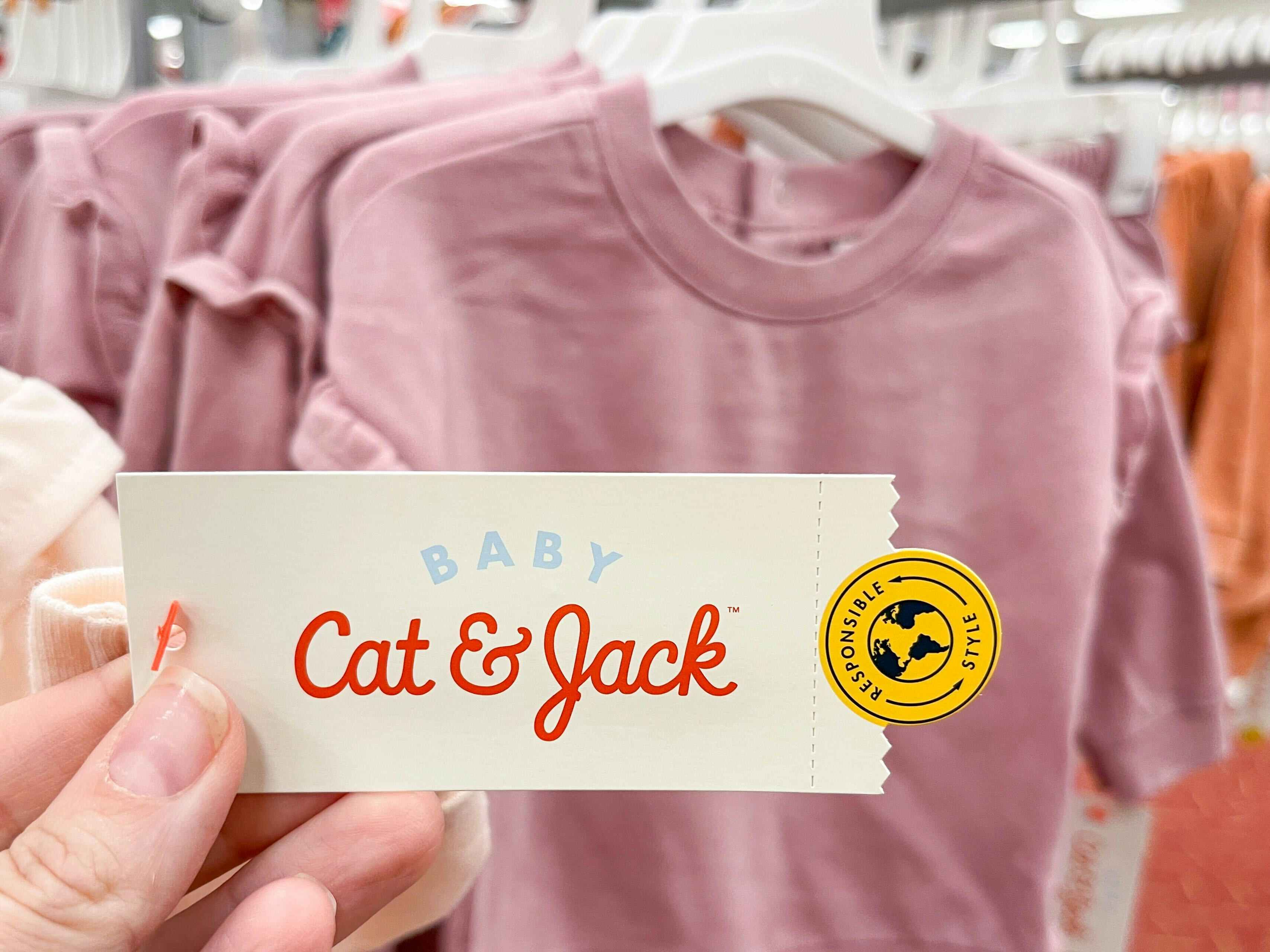 A Baby Cat & Jack tag held out by hand with infant clothes hanging in the back ground.