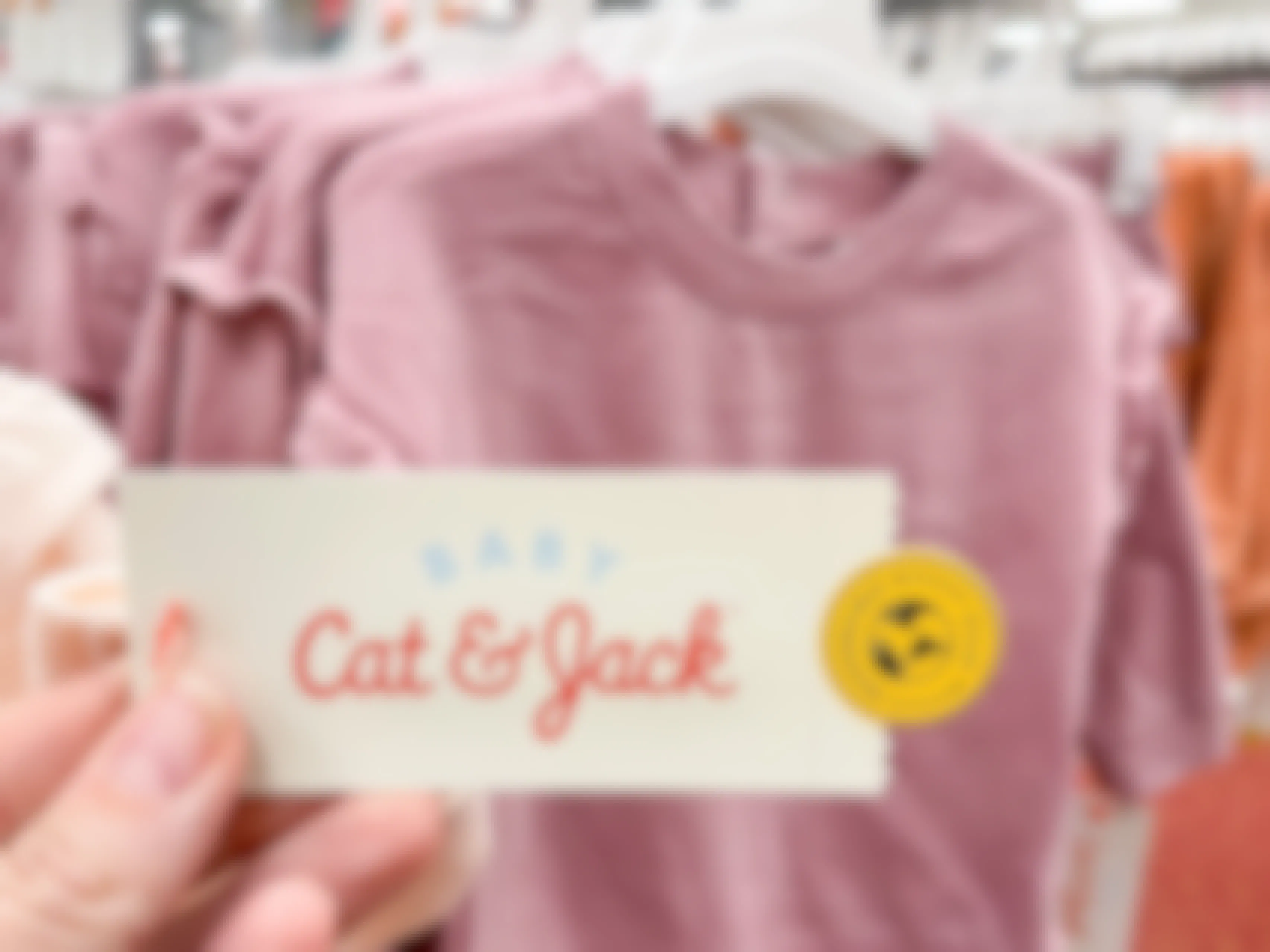 A Baby Cat & Jack tag held out by hand with infant clothes hanging in the back ground.