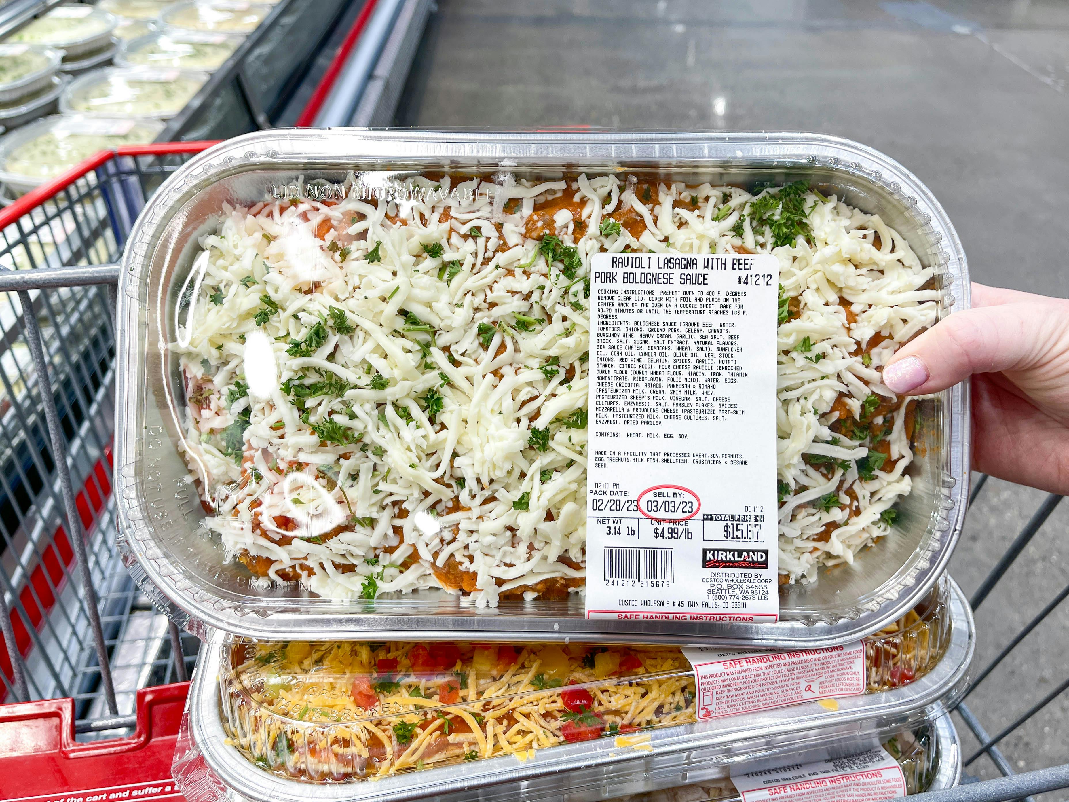 a person holding up a costco prepared meal in front of cart 