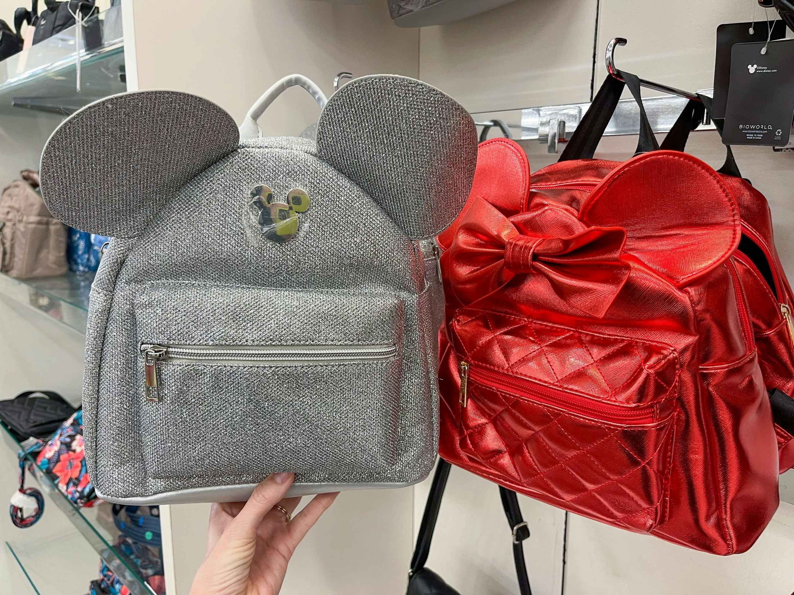 A variety of Disney backpacks. One held out by hand next to another one hanging from a store rack.