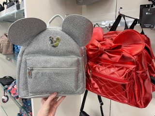 A variety of Disney backpacks. One held out by hand next to another one hanging from a store rack.