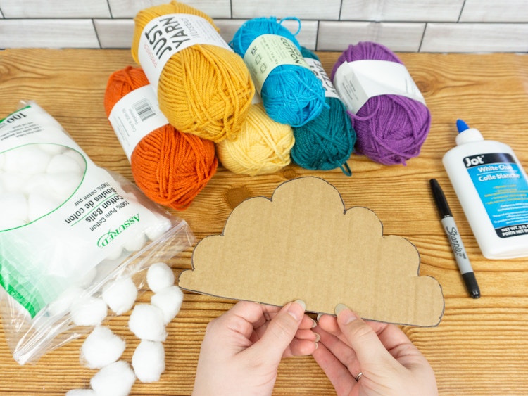 Someone holding a piece of cardboard cut out into a cloud shape in front of supplies for a DIY rainbow cloud decoration including different colored yarn, cotton balls, and glue