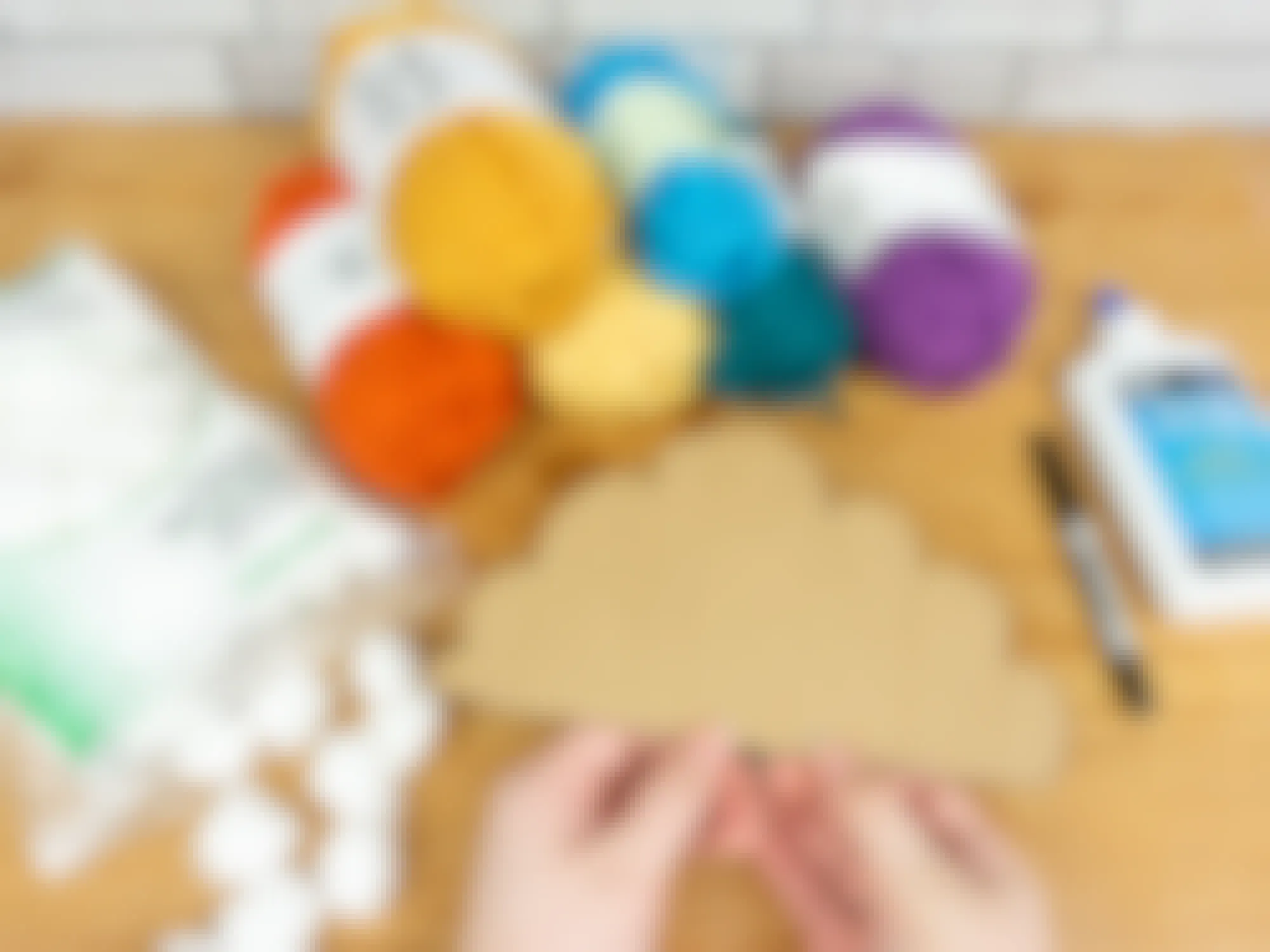 Someone holding a piece of cardboard cut out into a cloud shape in front of supplies for a DIY rainbow cloud decoration including different colored yarn, cotton balls, and glue