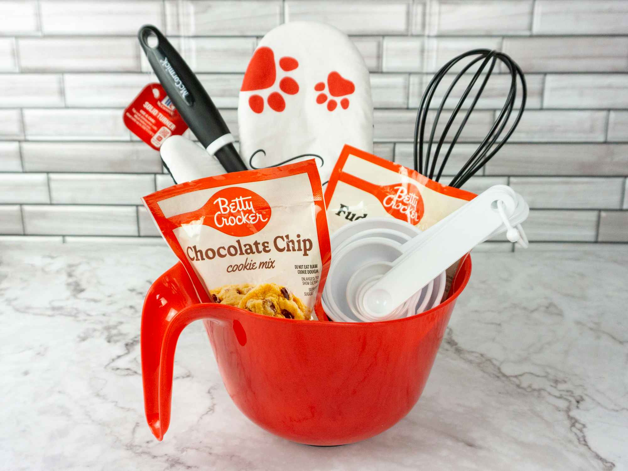 A baking themed gift basket with cookie mix, brownie mix, a whisk, an oven mitt, measuring cups, spatula, and a mixing bowl as the basket