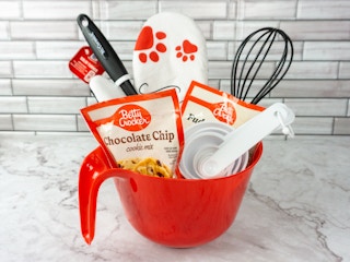 A baking themed gift basket with cookie mix, brownie mix, a whisk, an oven mitt, measuring cups, spatula, and a mixing bowl as the basket