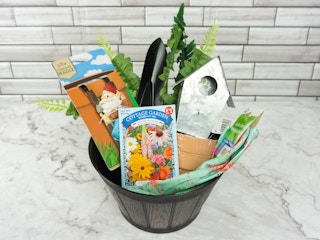 A gardening gift basket with a planter as the basket, and fake plants, gardening gloves, a trowel, some small pots, a little bird house, some seeds, and a gnome planter hugger inside