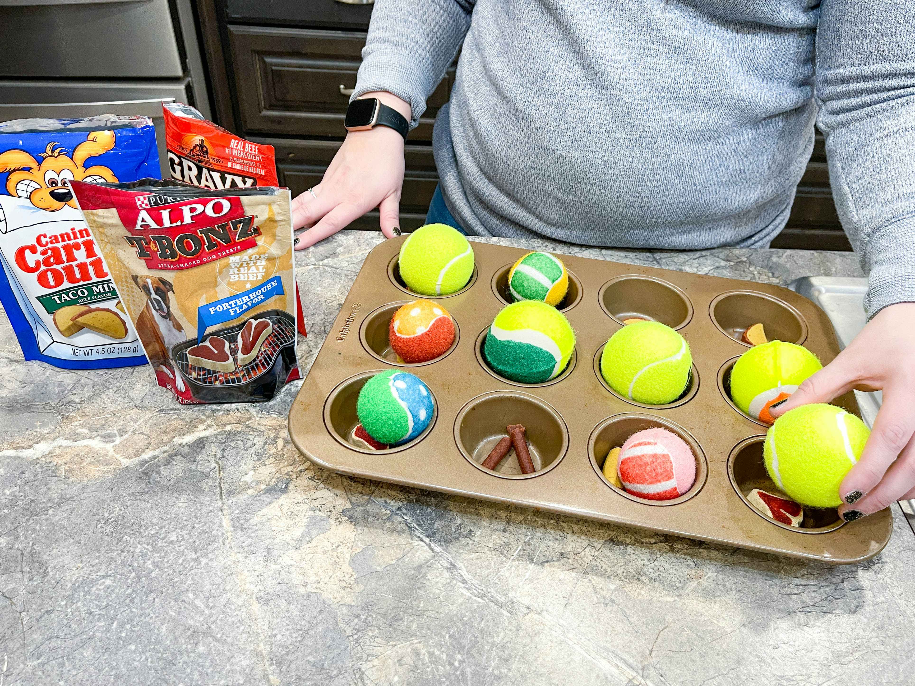 a person putting treats and tennis balls in a muffin tin for a dog toy 