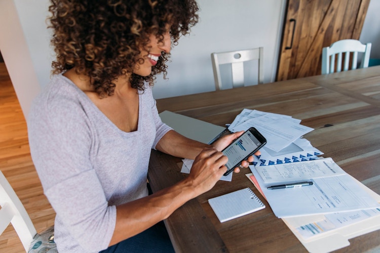A person looking at their phone with financial papers on the table in front of them