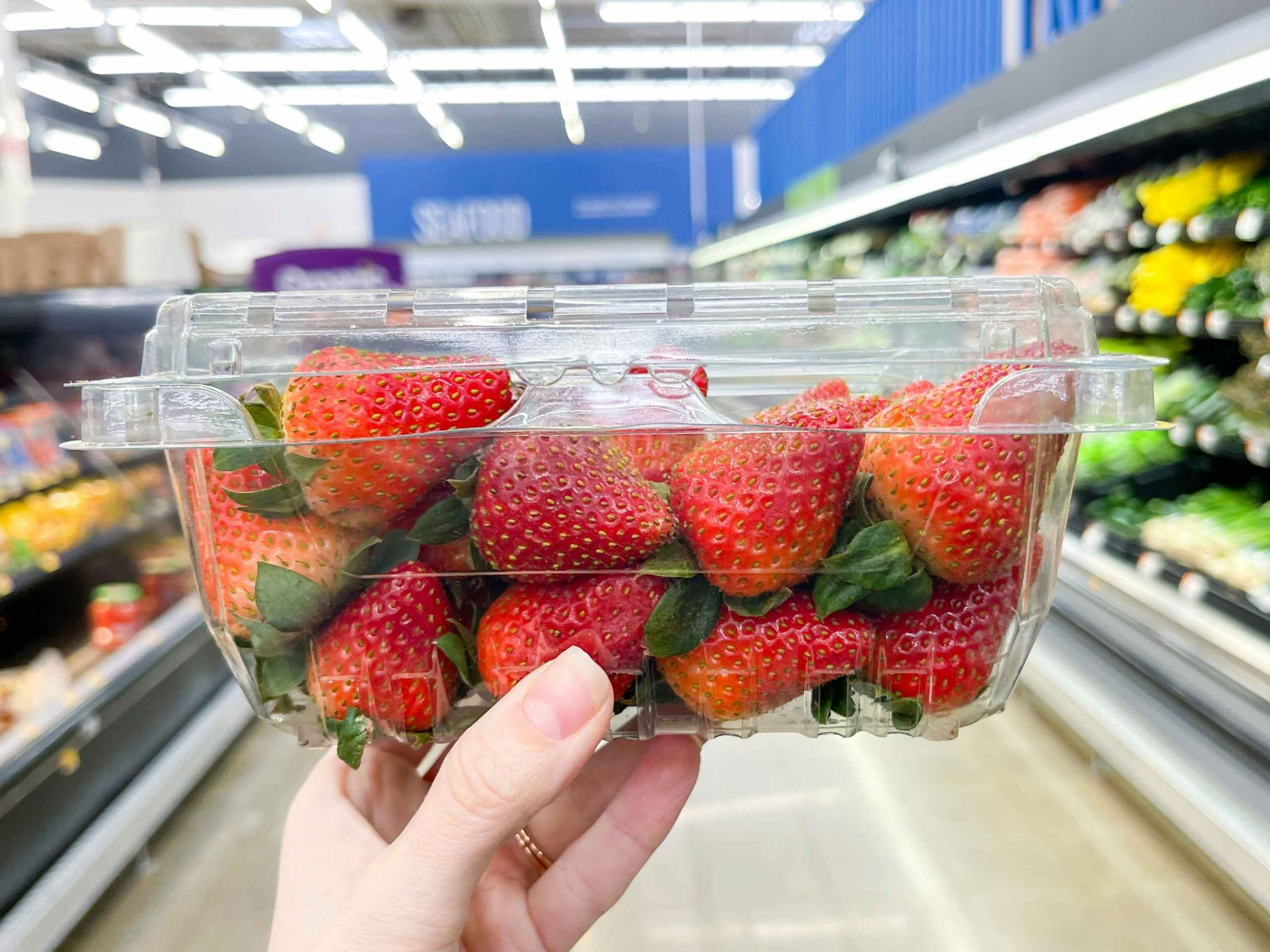 A box of strawberries held out by hand in front of a store aisle.